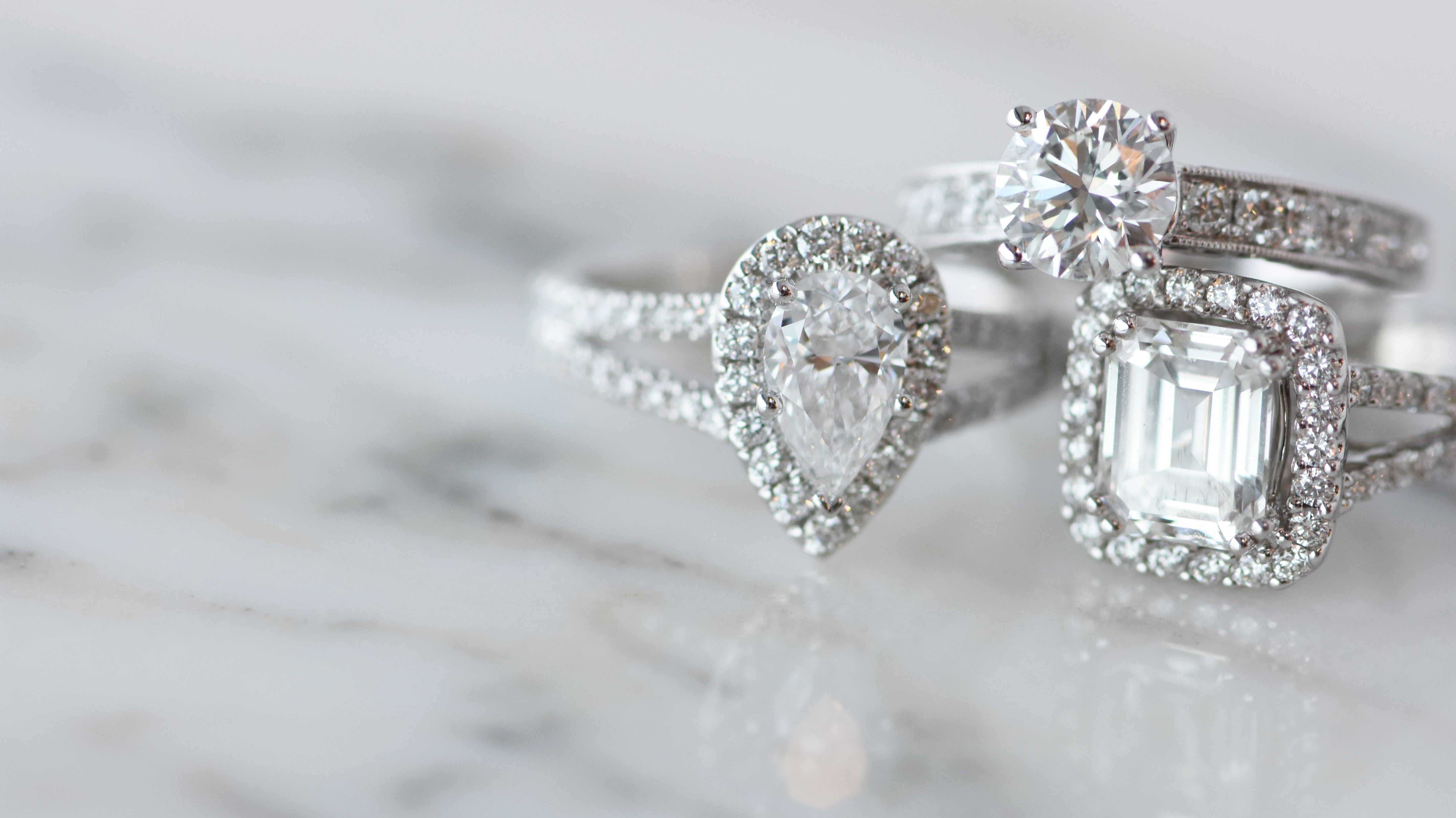 Three diamond engagement rings with halo settings—pear, round, and emerald cuts—set in white metal bands, displayed on a soft marble surface.