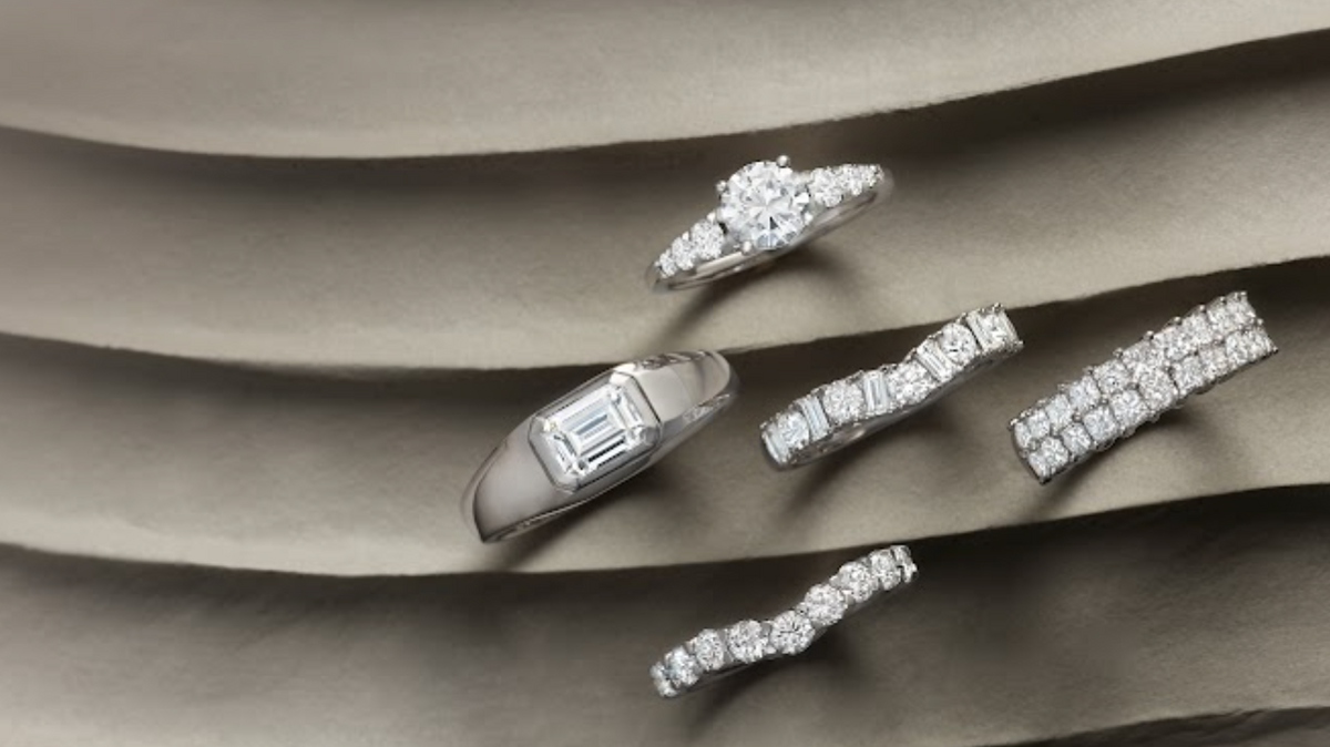 Five diamond rings arranged on layered gray fabric, featuring round, emerald-cut, channel-set, and pavé diamond designs.