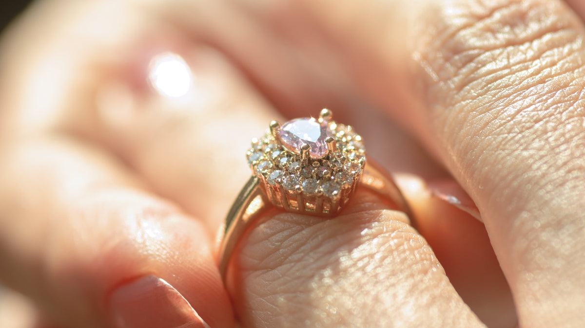Close-up of a hand holding a gold ring with a raised, halo-style setting featuring a pale pink center stone surrounded by small sparkling stones.