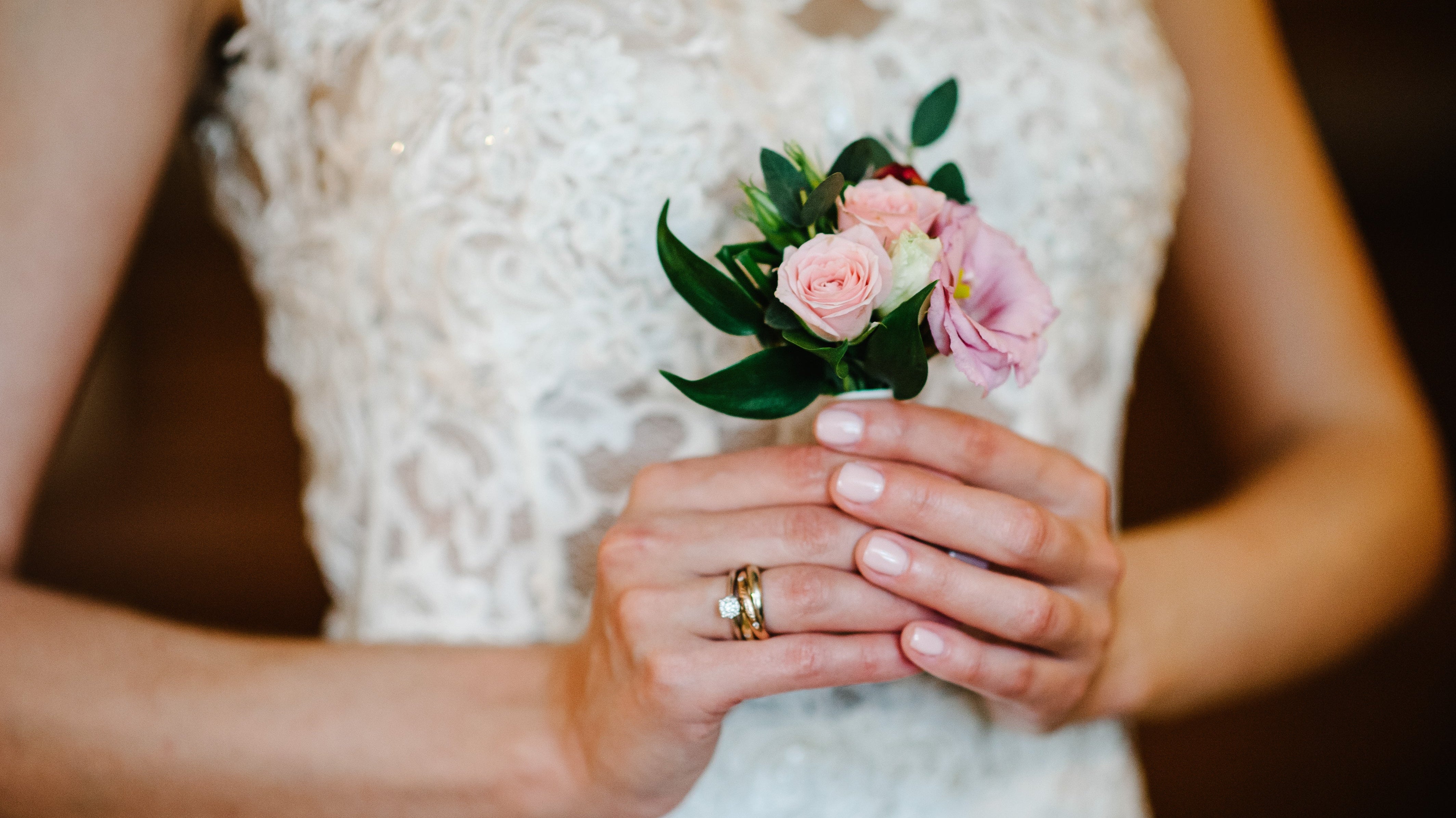 Bride holding a small floral bouquet, showing a diamond engagement ring stacked with a gold wedding band on her left hand during the wedding day.
