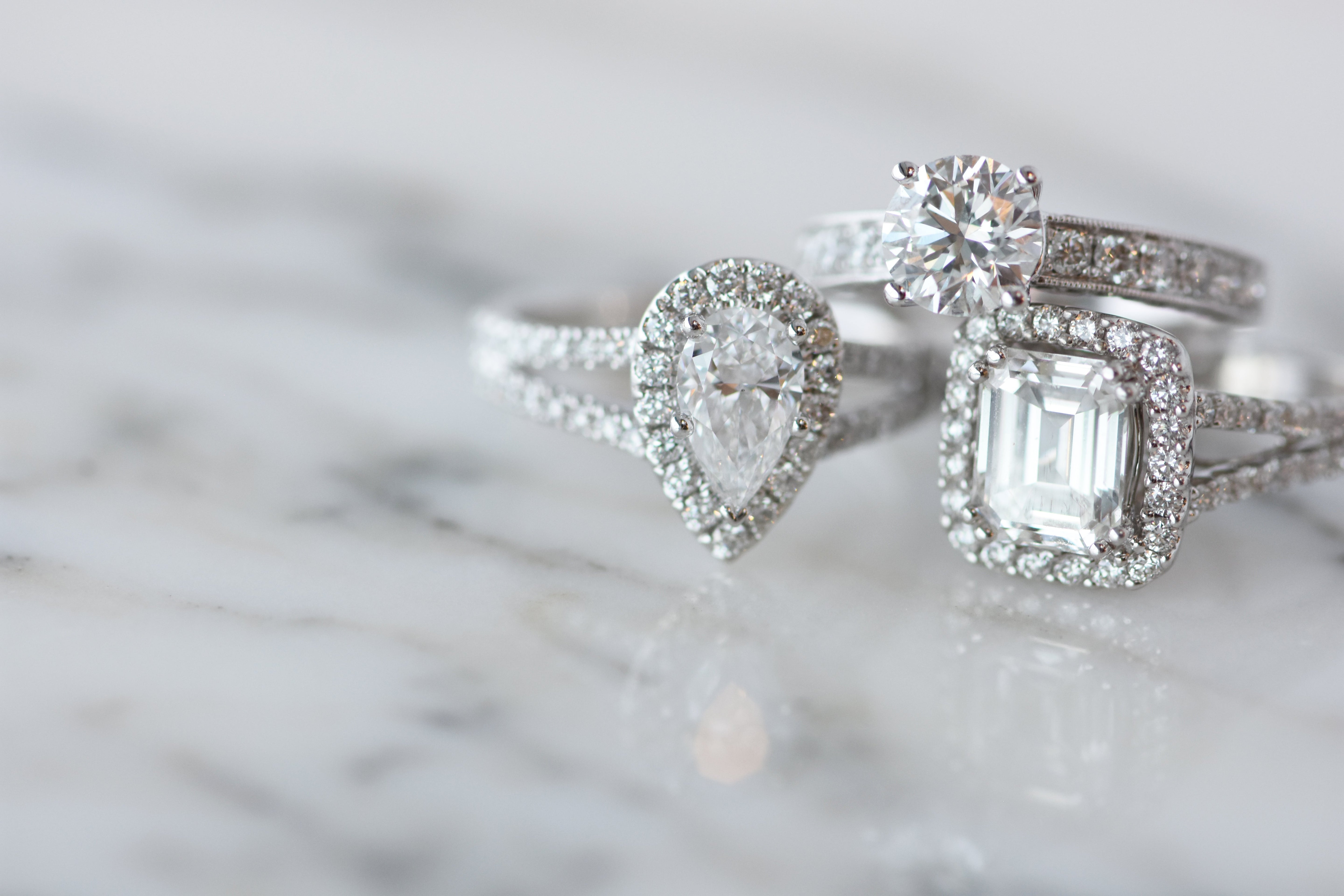 Three diamond engagement rings with halo settings—pear, round, and emerald cuts—set in white metal bands, displayed on a soft marble surface.