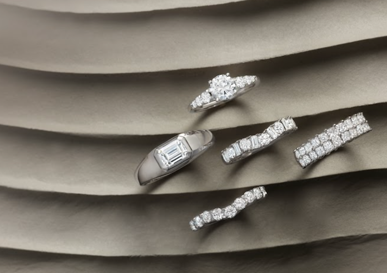 Five diamond rings arranged on layered gray fabric, featuring round, emerald-cut, channel-set, and pavé diamond designs.
