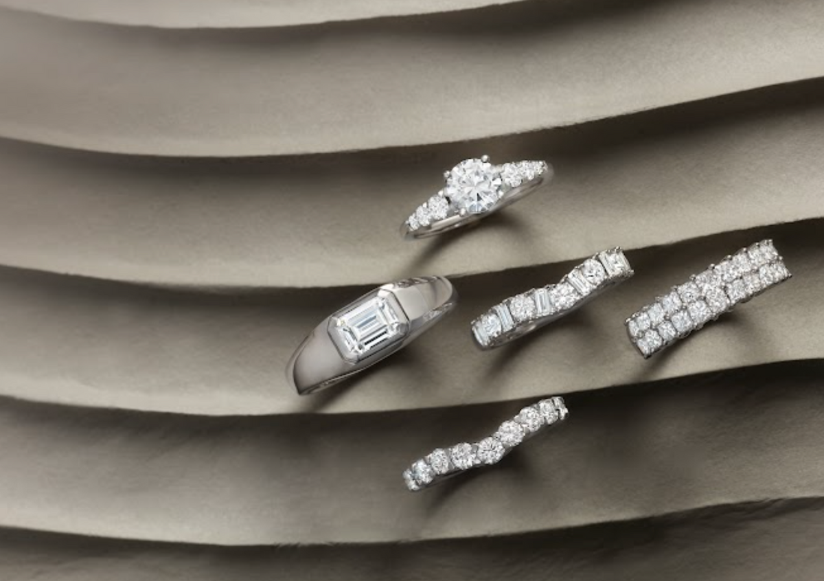 Five diamond rings arranged on layered gray fabric, featuring round, emerald-cut, channel-set, and pavé diamond designs.