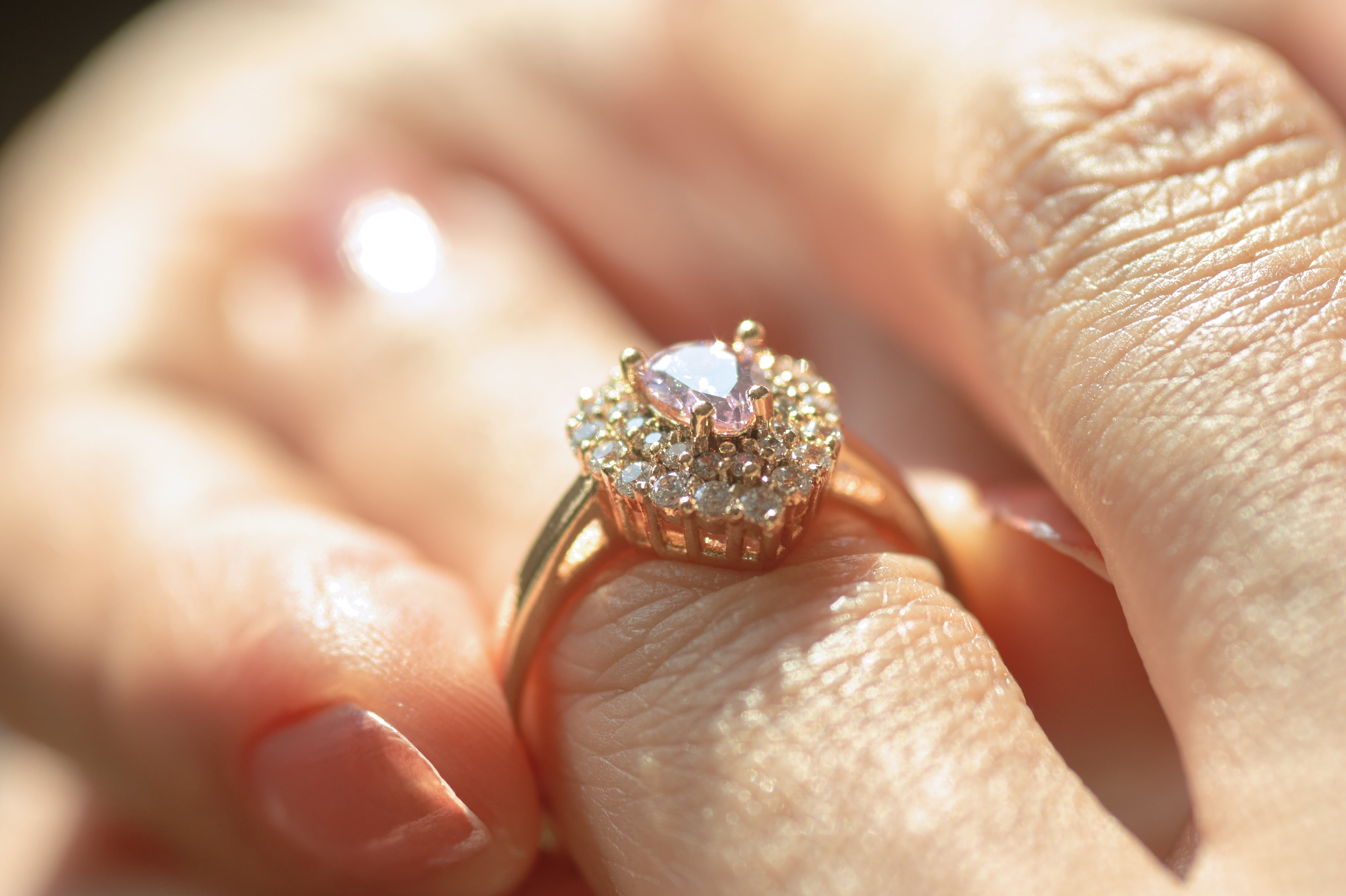 Close-up of a hand holding a gold ring with a raised, halo-style setting featuring a pale pink center stone surrounded by small sparkling stones.