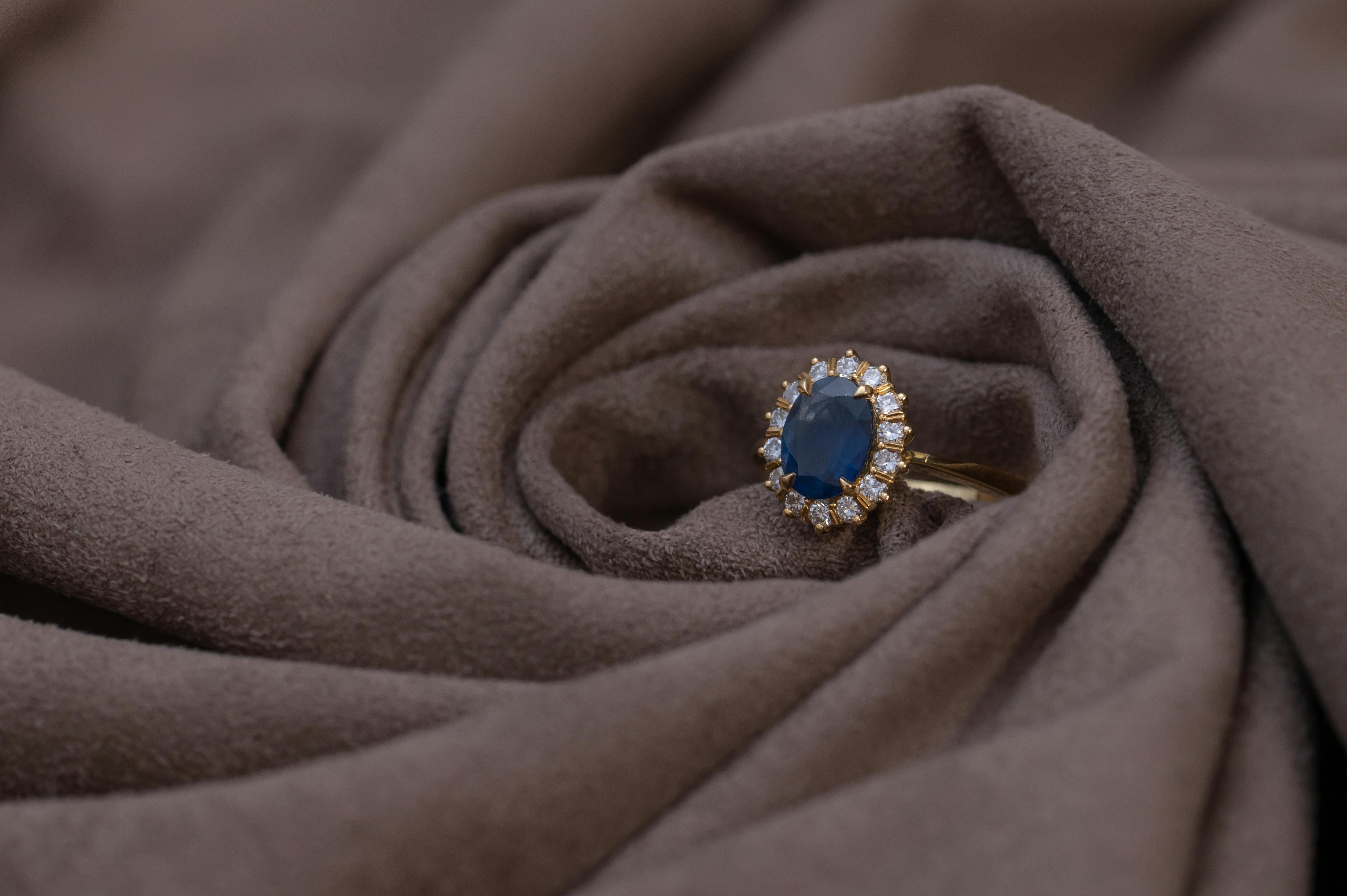 Gold statement ring featuring an oval blue sapphire surrounded by a halo of diamonds, displayed elegantly on soft brown fabric.