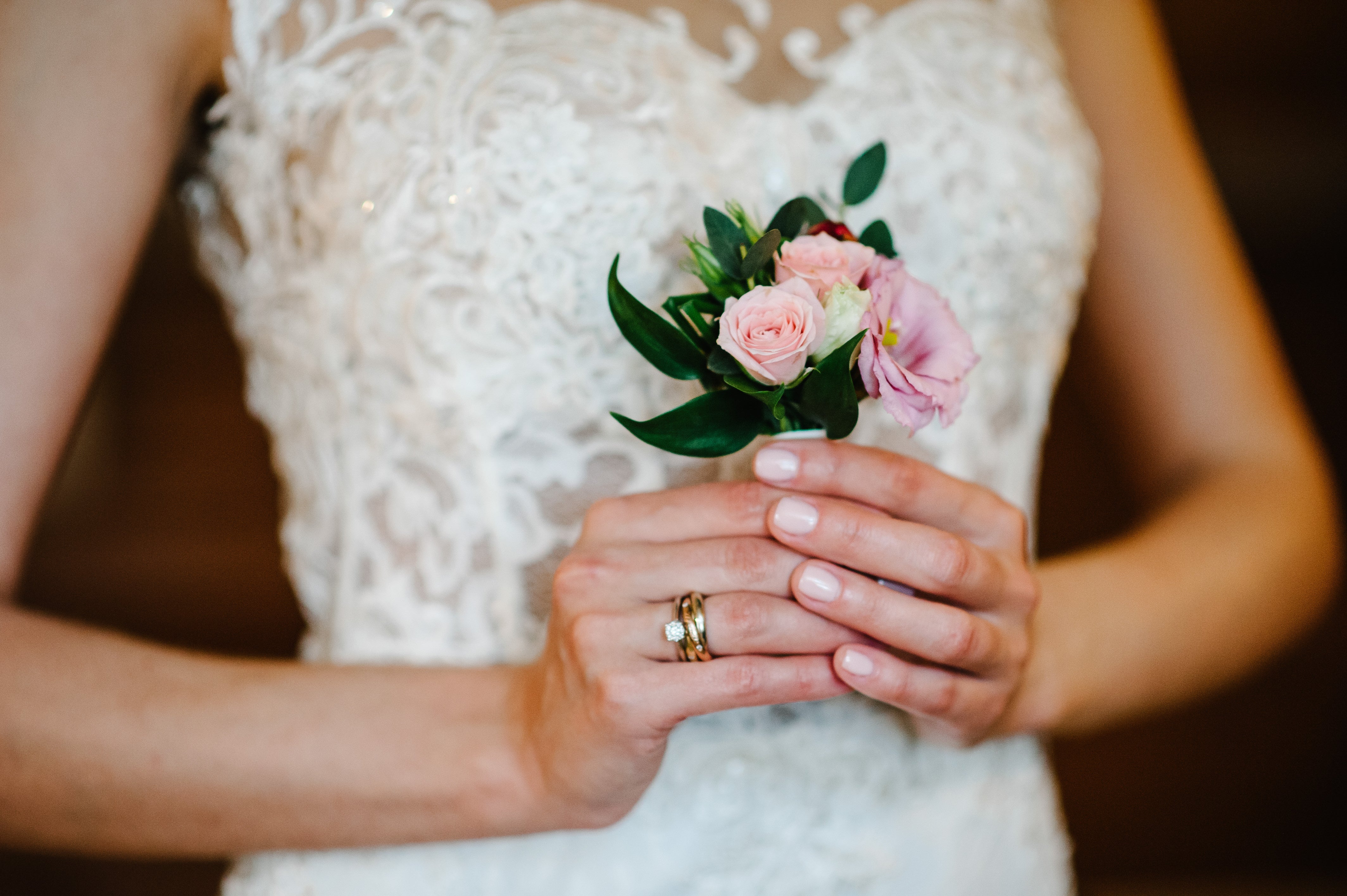 Bride holding a small floral bouquet, showing a diamond engagement ring stacked with a gold wedding band on her left hand during the wedding day.