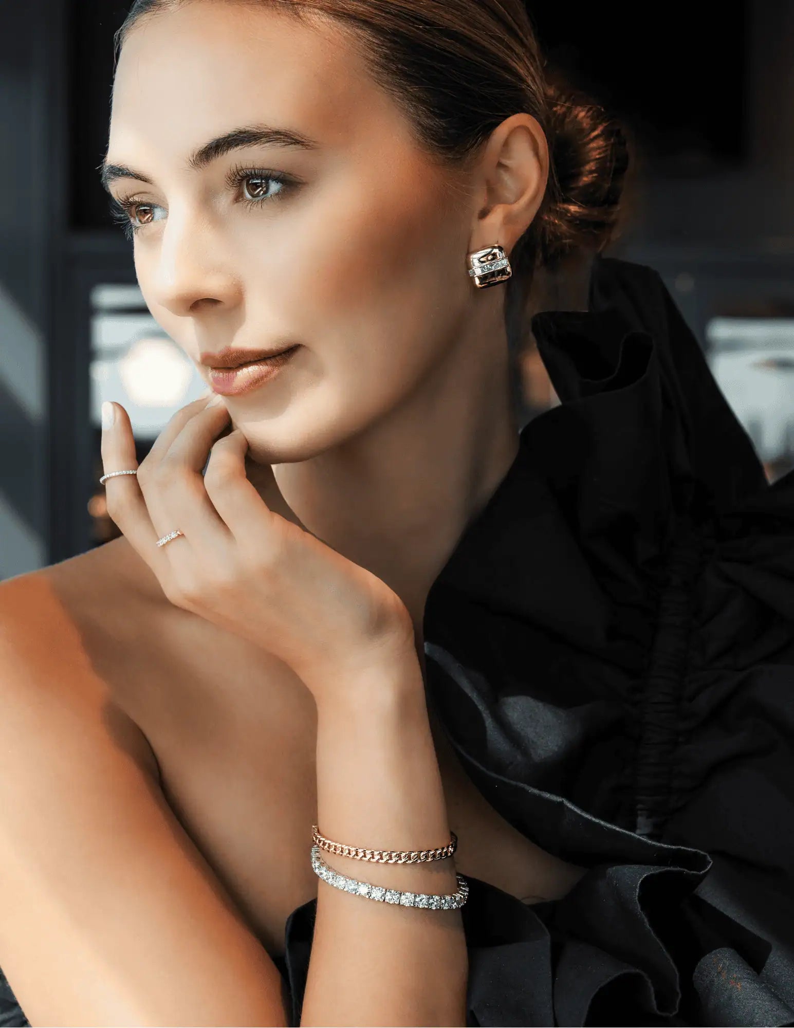 Elegant portrait of a woman wearing gold earrings, a diamond bracelet, and delicate rings, styled with a black ruffled garment and warm, soft lighting to highlight the jewelry.