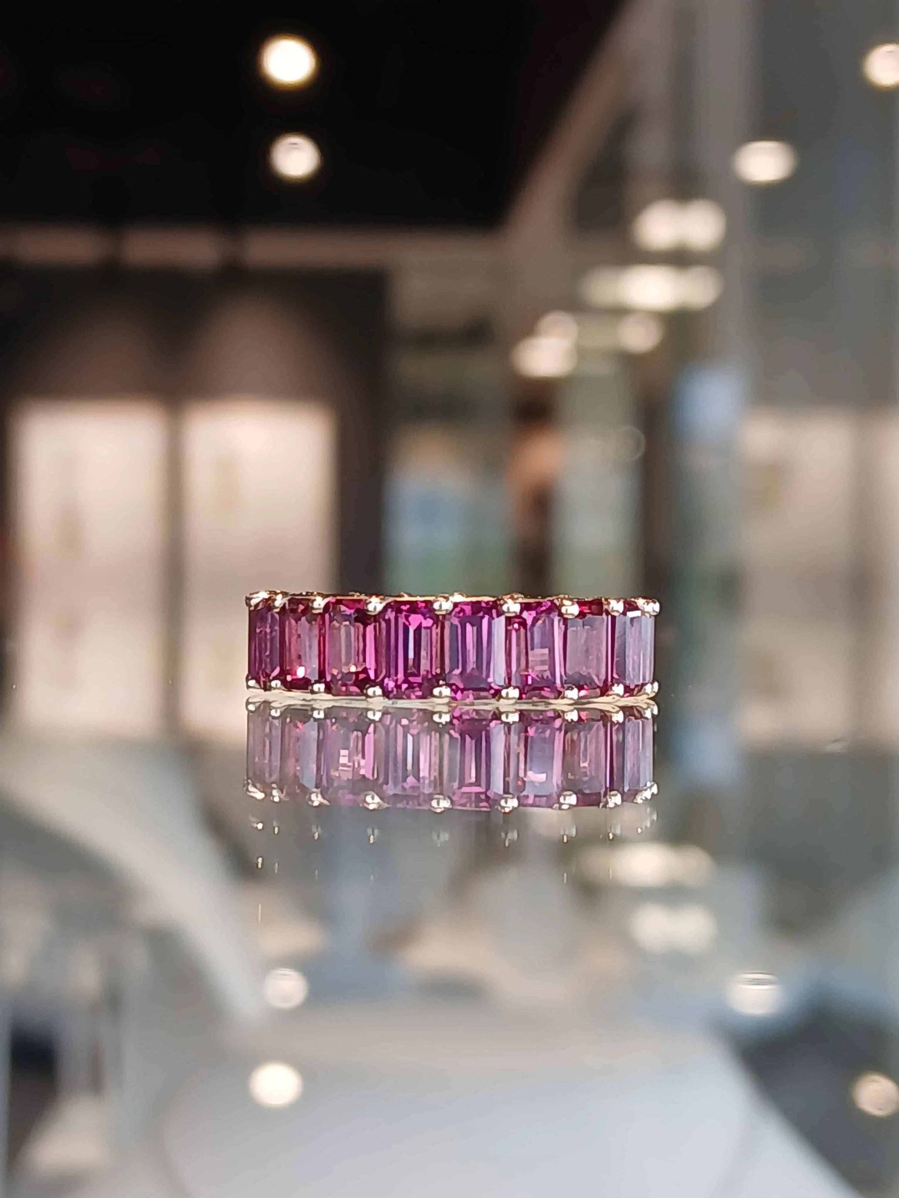 Pink baguette-cut gemstone eternity band set in yellow gold, displayed on a reflective surface with soft showroom lighting in the background.