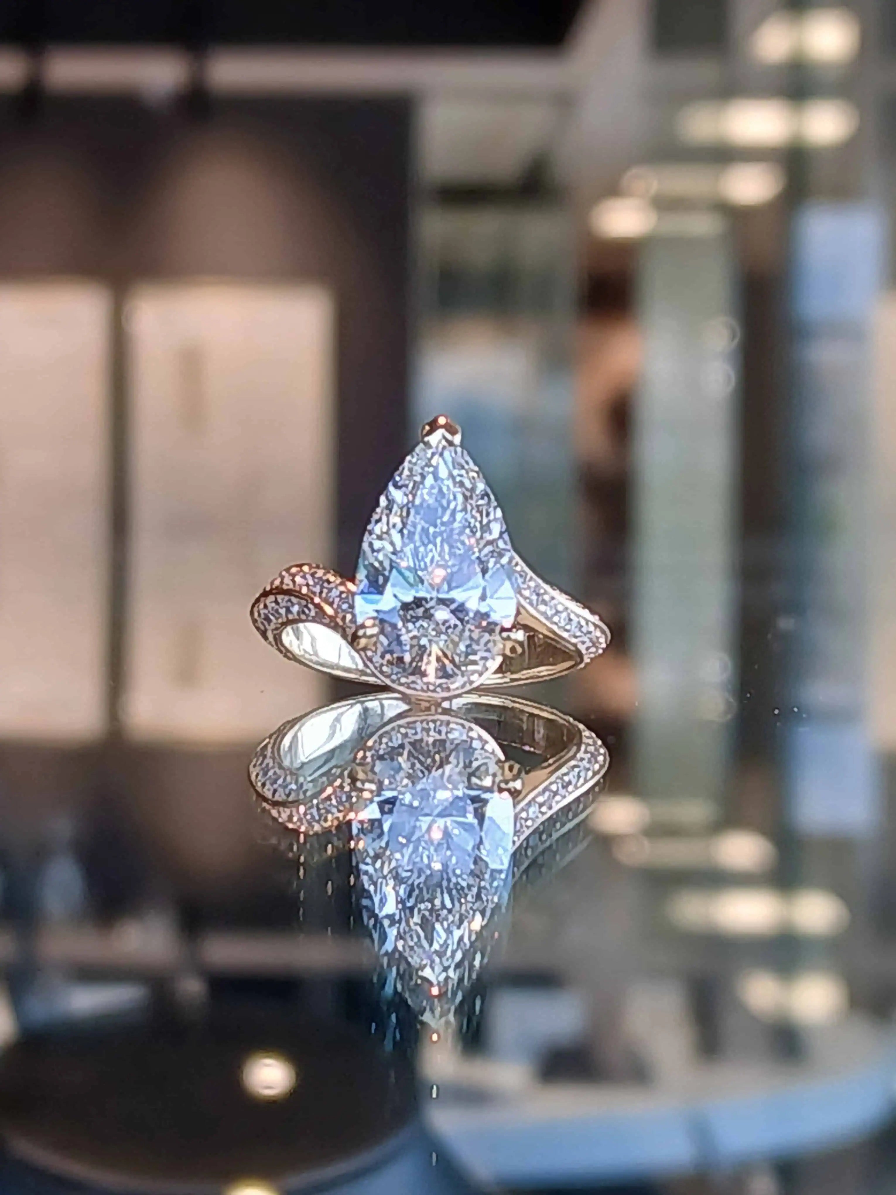 Pear-shaped diamond engagement ring with a pavé-set split shank in yellow gold, displayed on a reflective surface with a softly blurred jewelry showroom background.