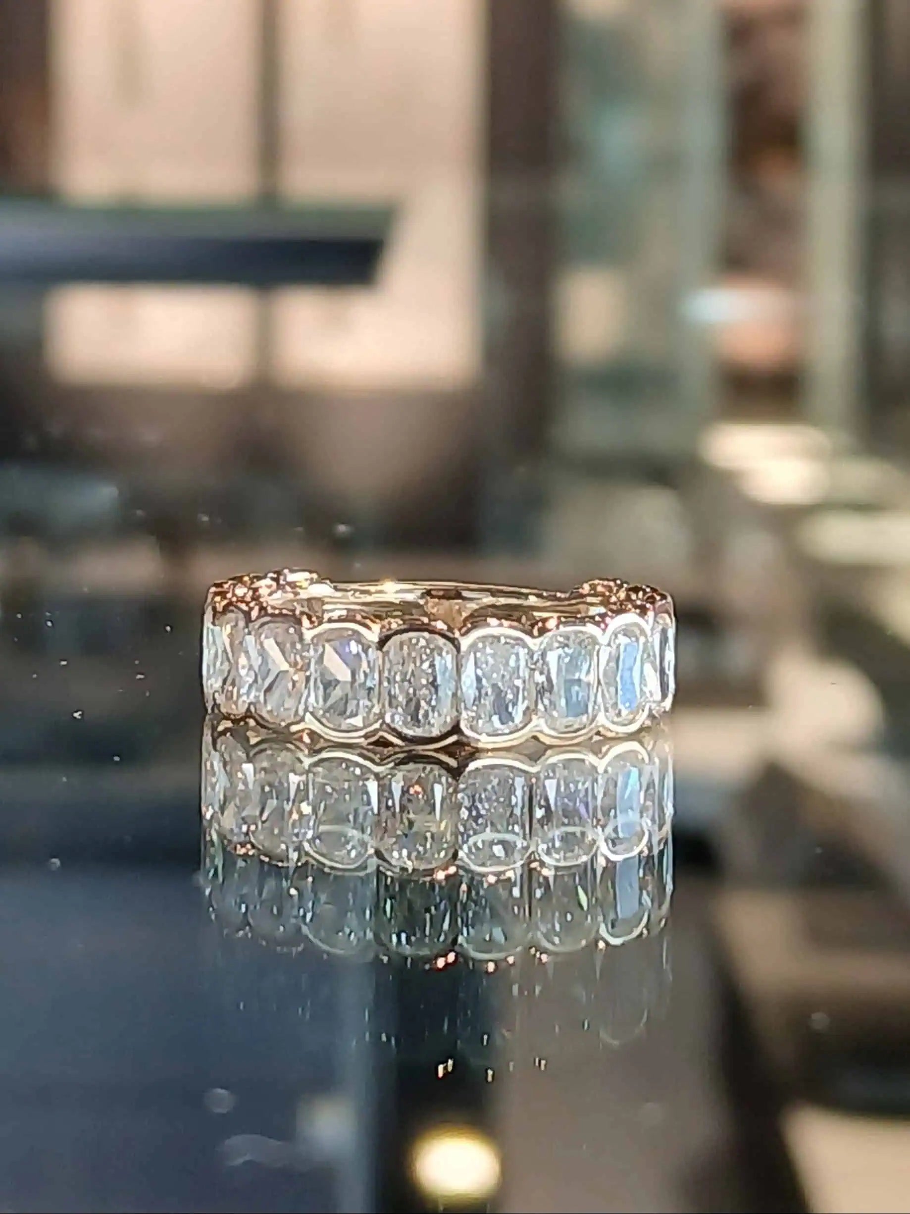 Diamond eternity band featuring a row of oval-cut diamonds set in yellow gold, displayed on a reflective surface with a softly blurred showroom background.
