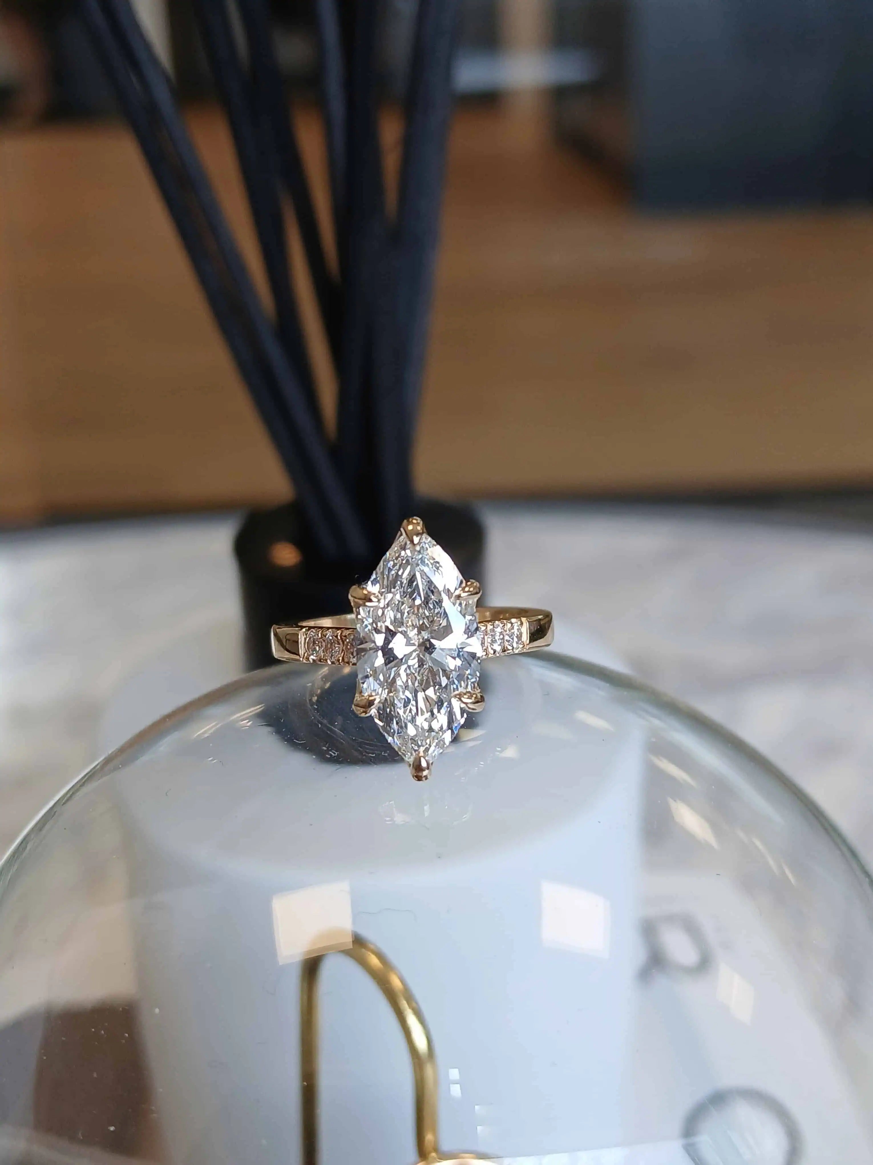 Close-up of a marquise-cut diamond engagement ring set in yellow gold with pavé band details, displayed on a glass dome with soft ambient lighting.