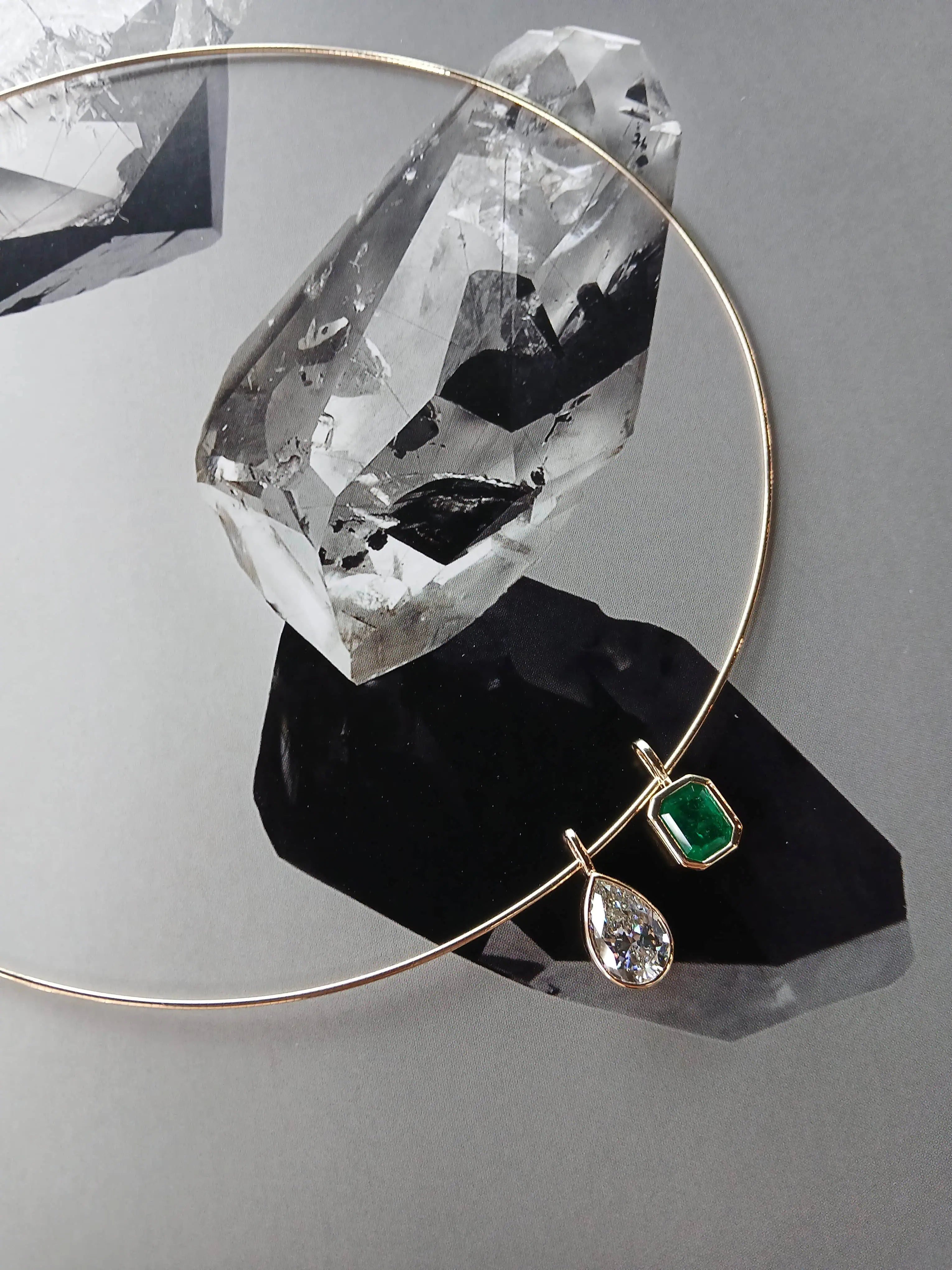 Minimalist gold necklace featuring a pear-shaped diamond pendant and an emerald-cut green gemstone pendant, styled against a crystal graphic background.