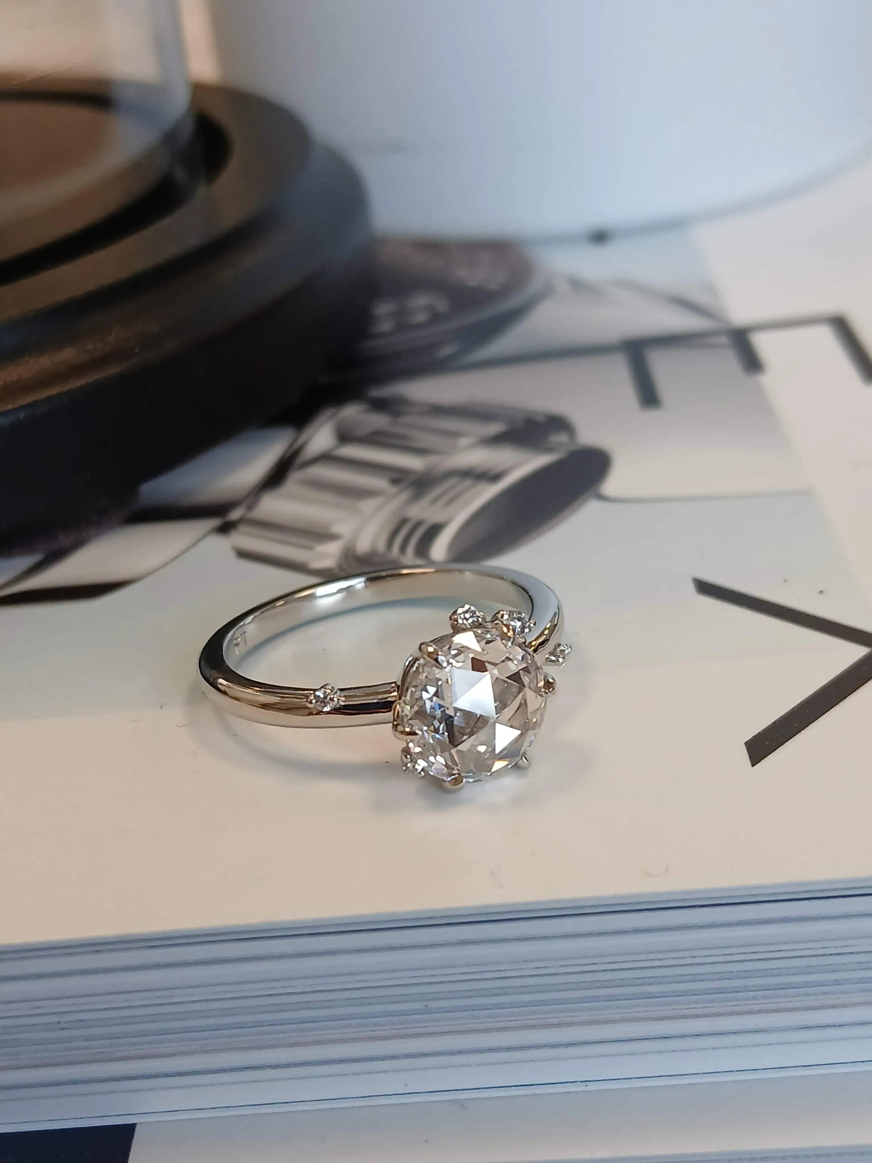 Close-up of a diamond engagement ring with an oval-cut center stone and delicate side diamonds resting on an open book beside a decorative clock, highlighting elegant fine jewelry styling.