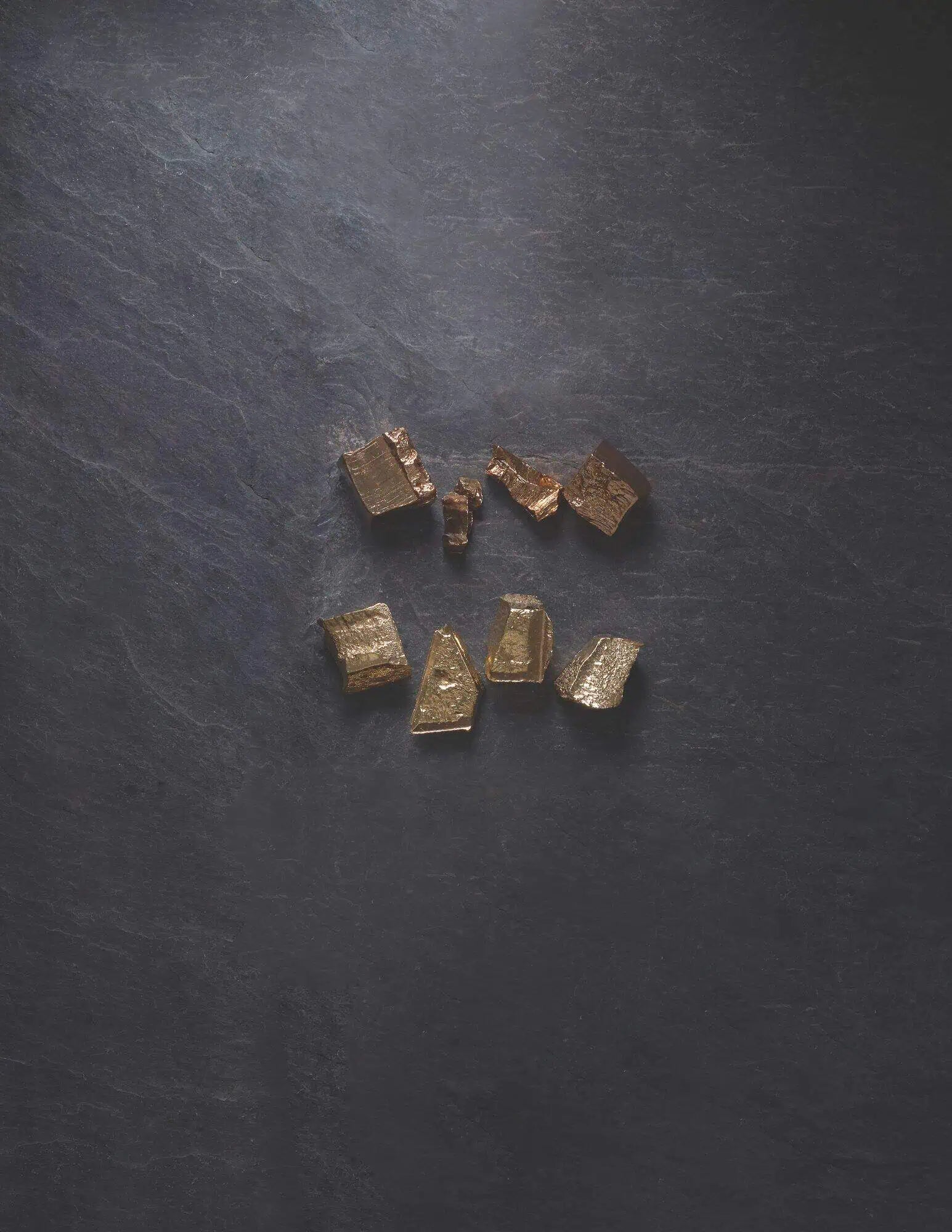 Raw gold nuggets displayed on a dark textured surface, representing natural precious metals used in fine jewelry crafting.