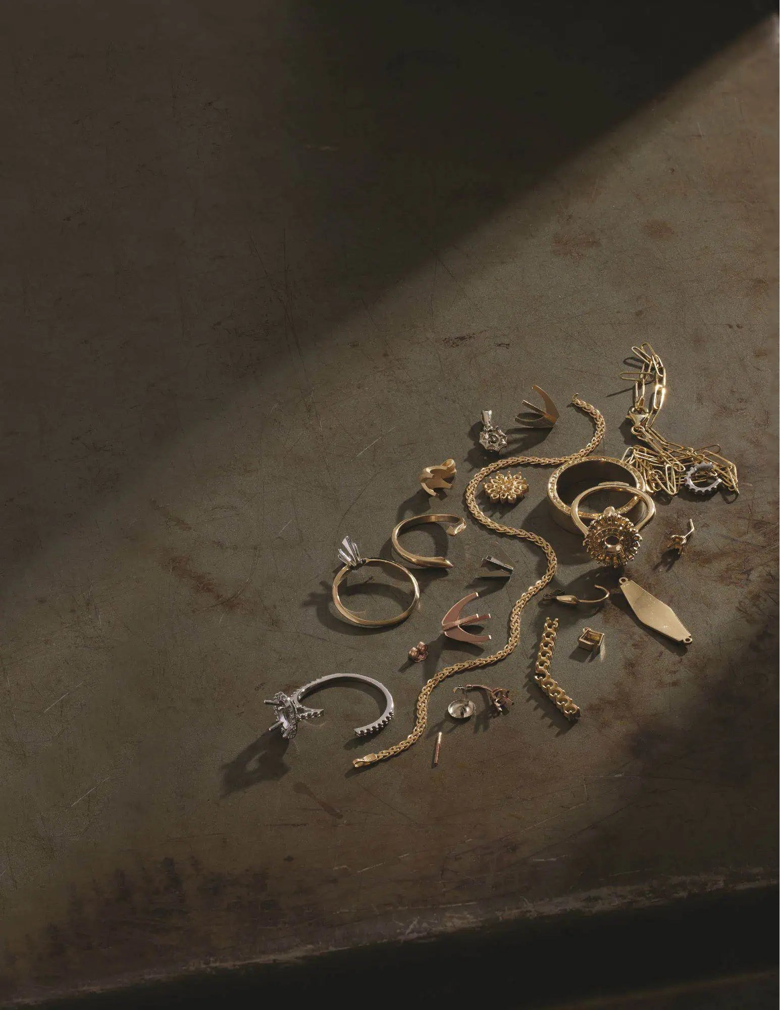 Assorted gold and silver jewelry components, ring settings, chains, and clasps arranged on a textured metal work surface, representing elements used in custom jewelry design and repair.