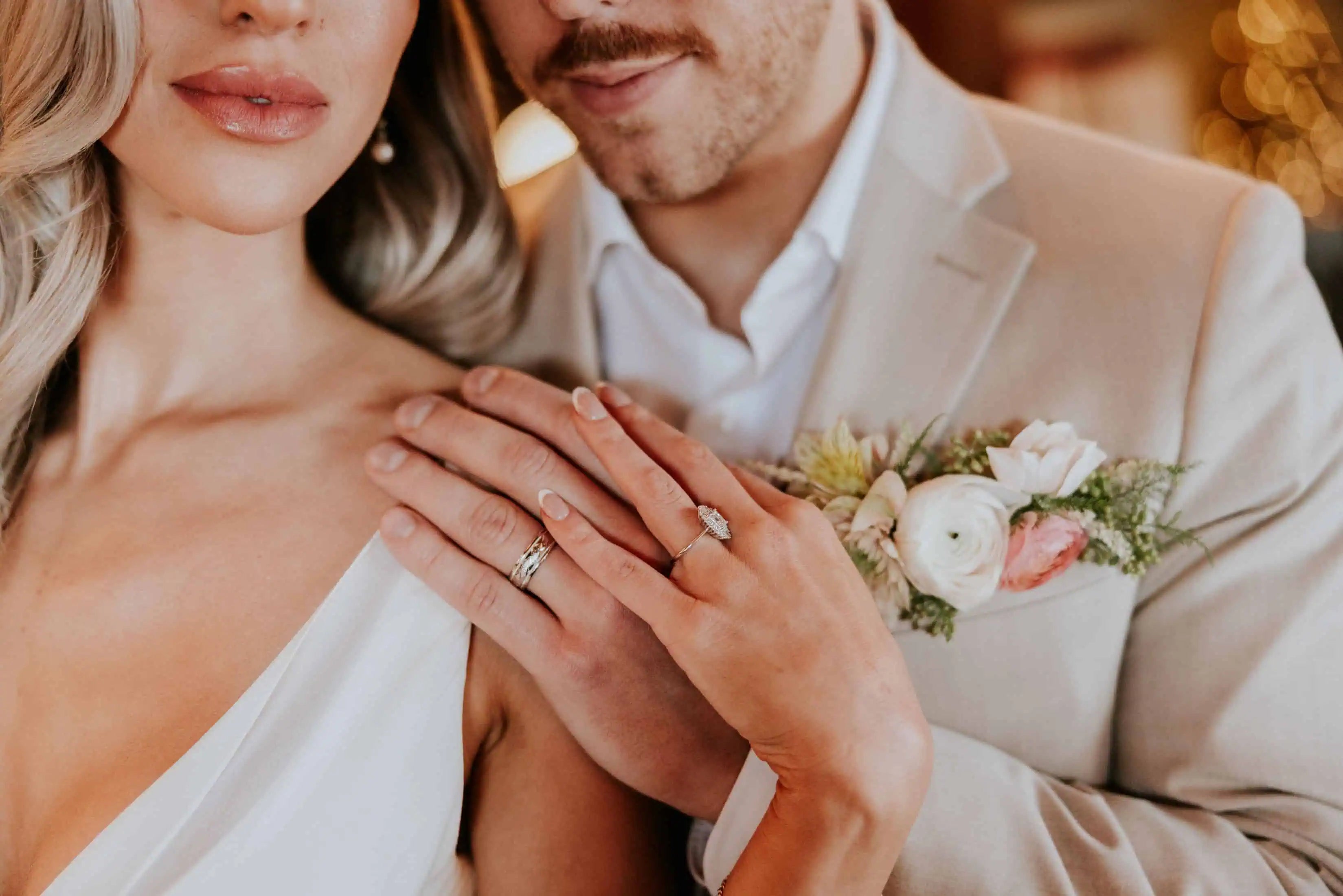 Close-up of a bride and groom with hands overlapping on the bride’s shoulder, showcasing diamond engagement and wedding rings beside a soft floral boutonniere.