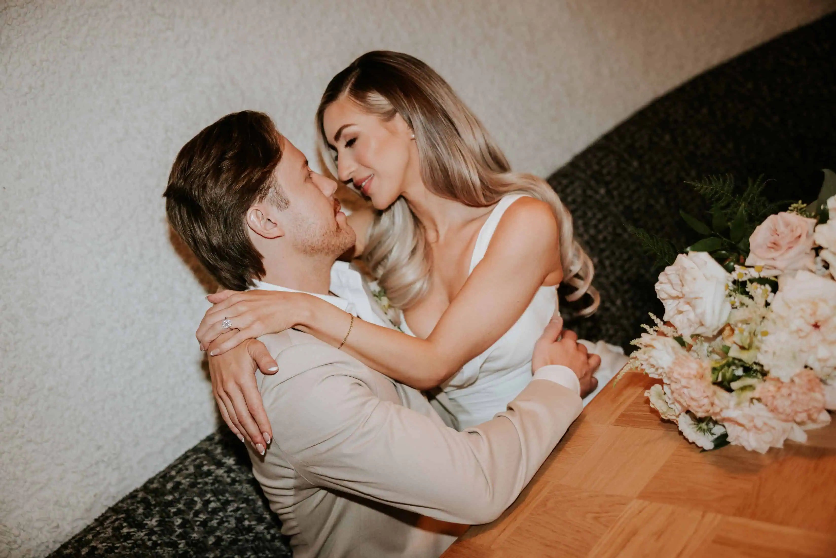 Romantic couple embracing at a table during a wedding celebration, with the bride wearing a diamond engagement ring and soft floral bouquet nearby.