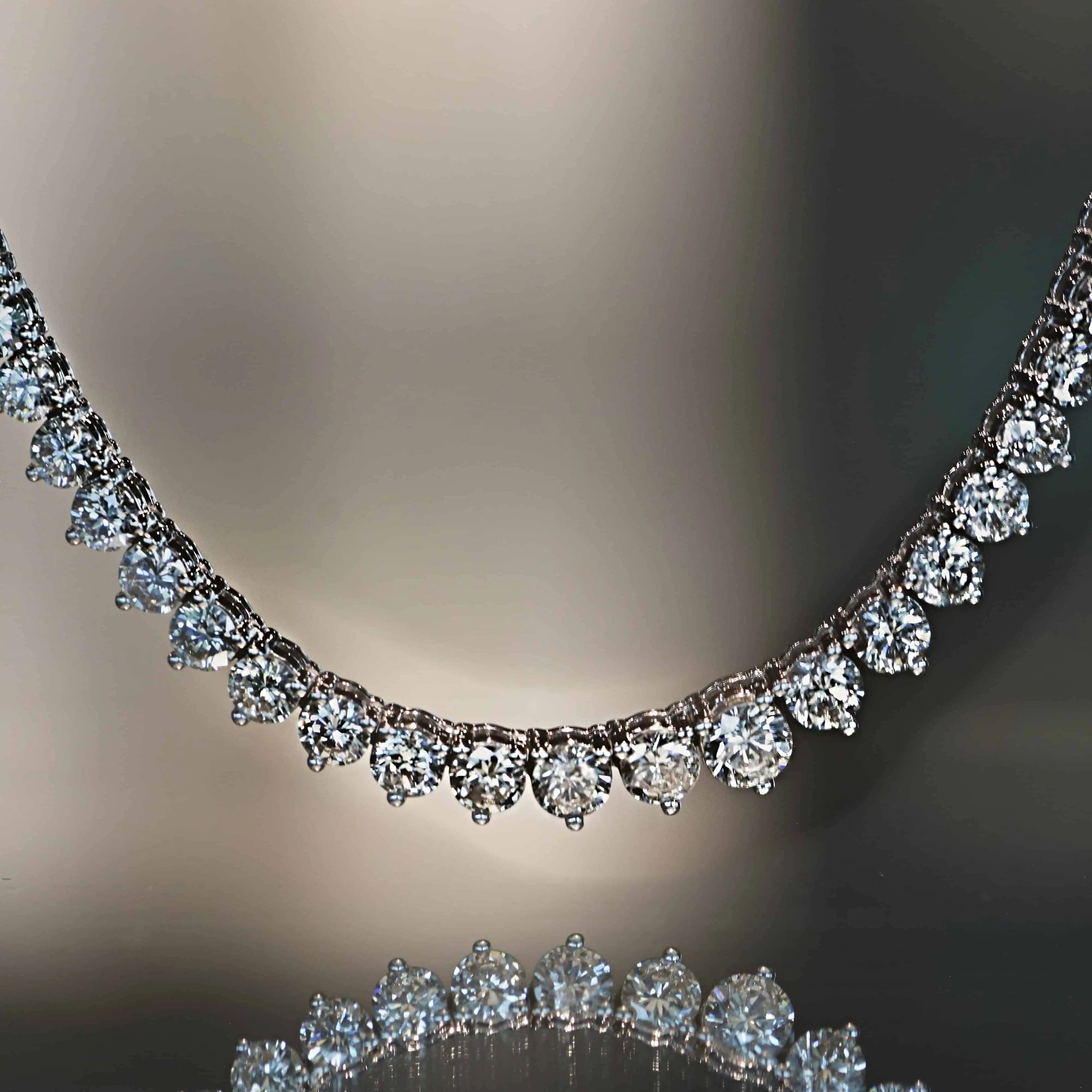 Close-up of a diamond necklace featuring a row of round brilliant diamonds in a graduated drop design, displayed on a reflective surface.