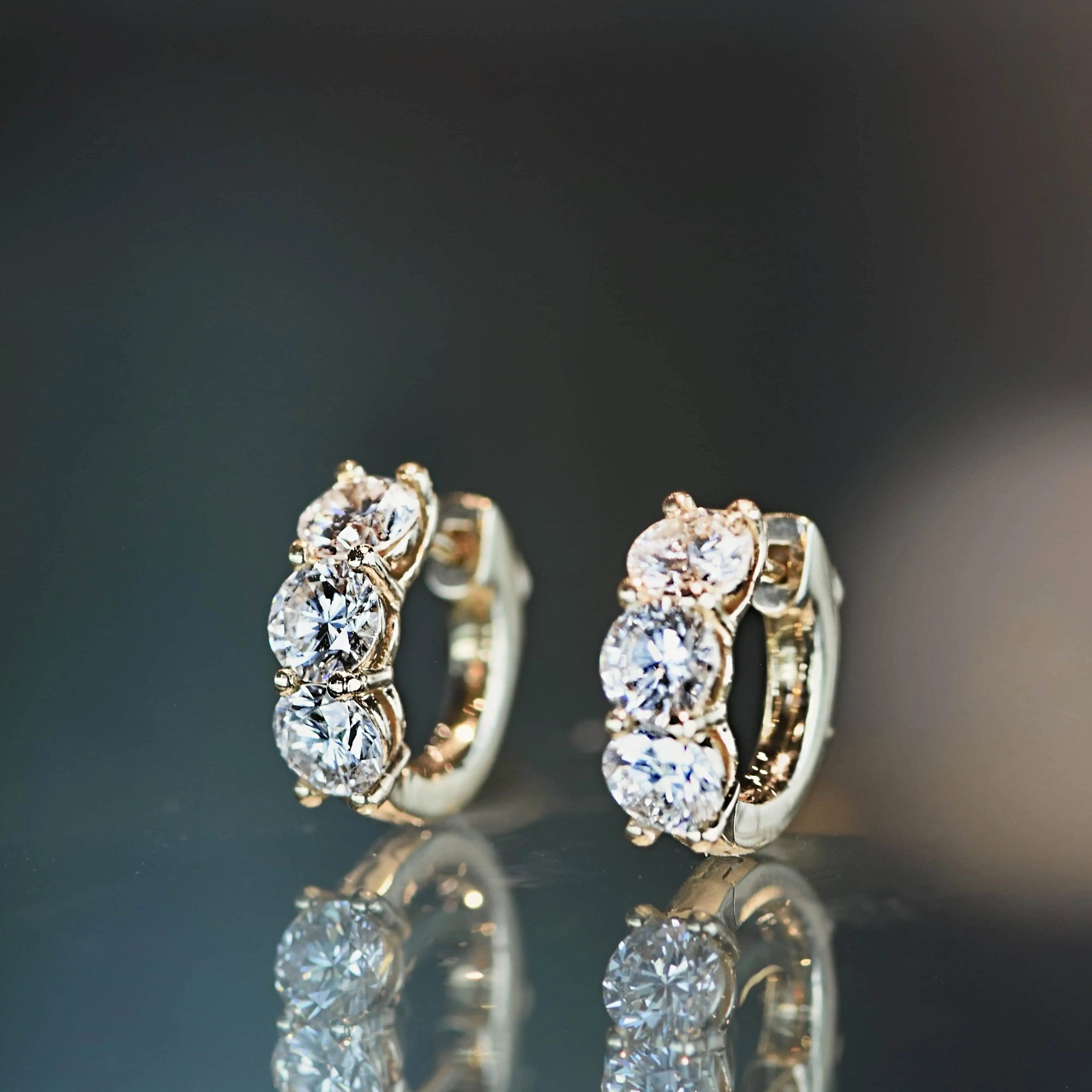 Pair of gold hoop earrings featuring three round brilliant diamonds each, displayed on a reflective surface with soft lighting.