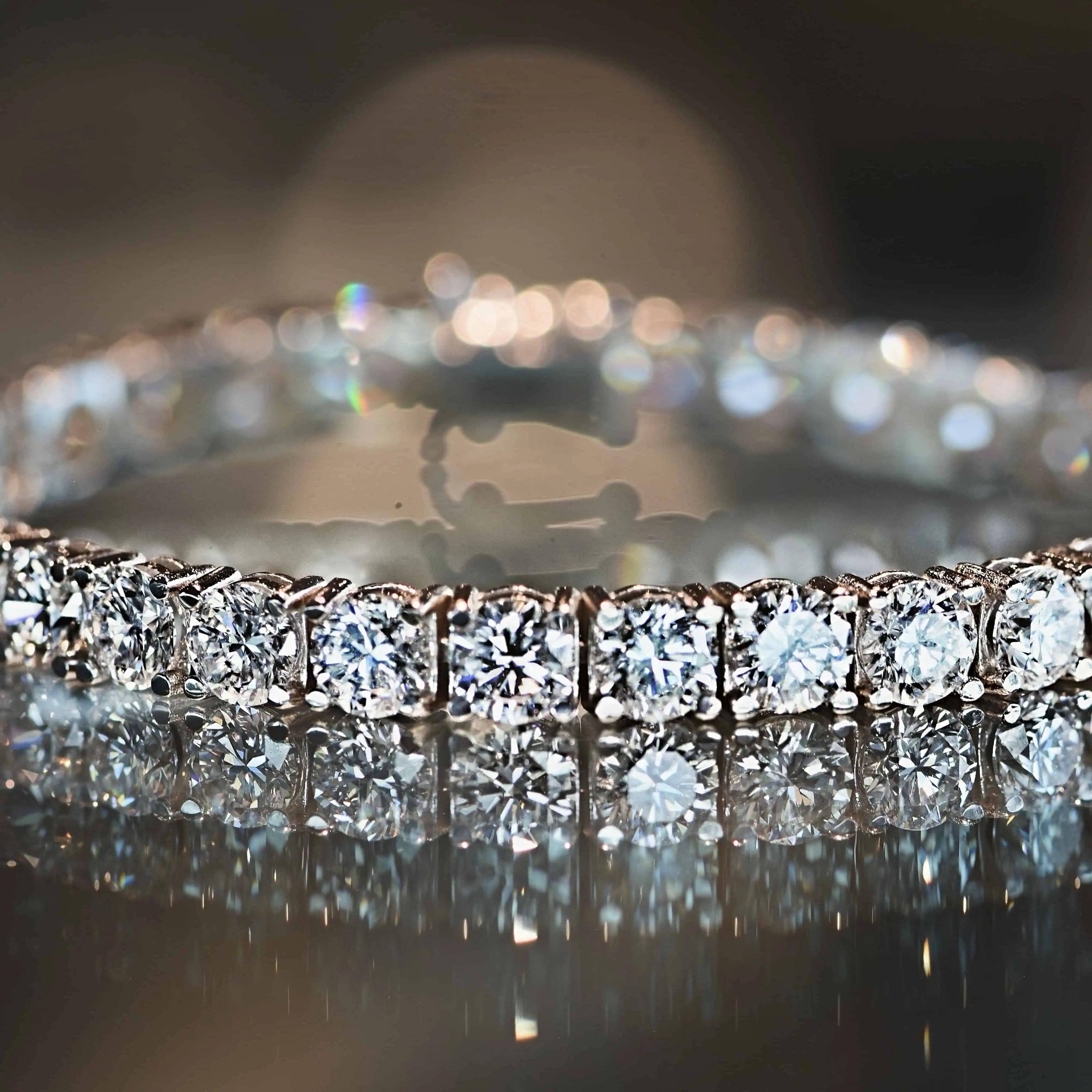 Close-up of a diamond tennis bracelet featuring round brilliant diamonds in a continuous setting, displayed on a reflective surface with soft bokeh lighting.