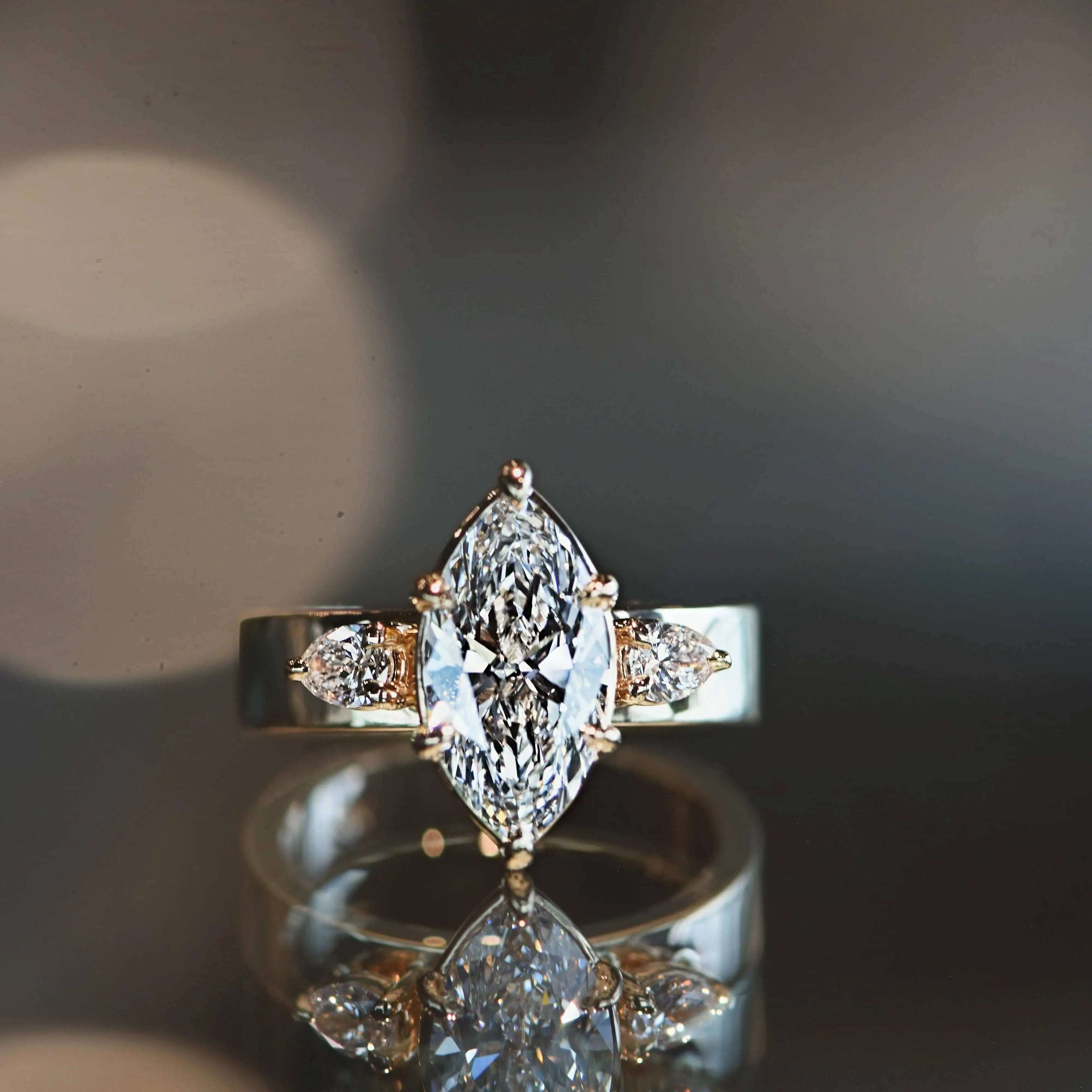 Close-up of a marquise-cut diamond engagement ring with pear-shaped side stones set in a polished band, reflected on a glossy surface.