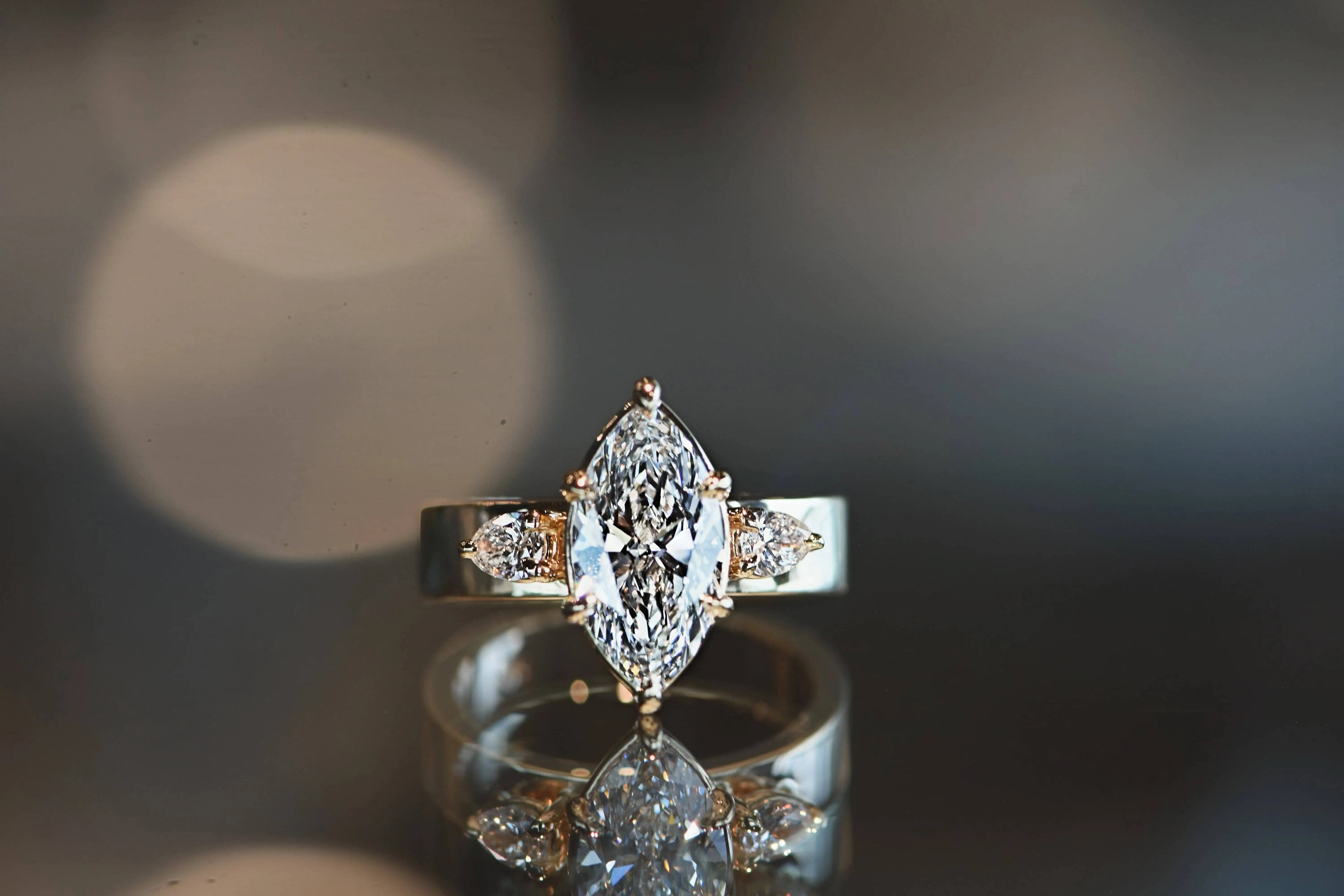 Marquise-cut diamond engagement ring with two pear-shaped side stones set in yellow gold, displayed on a reflective surface with soft ambient lighting.
