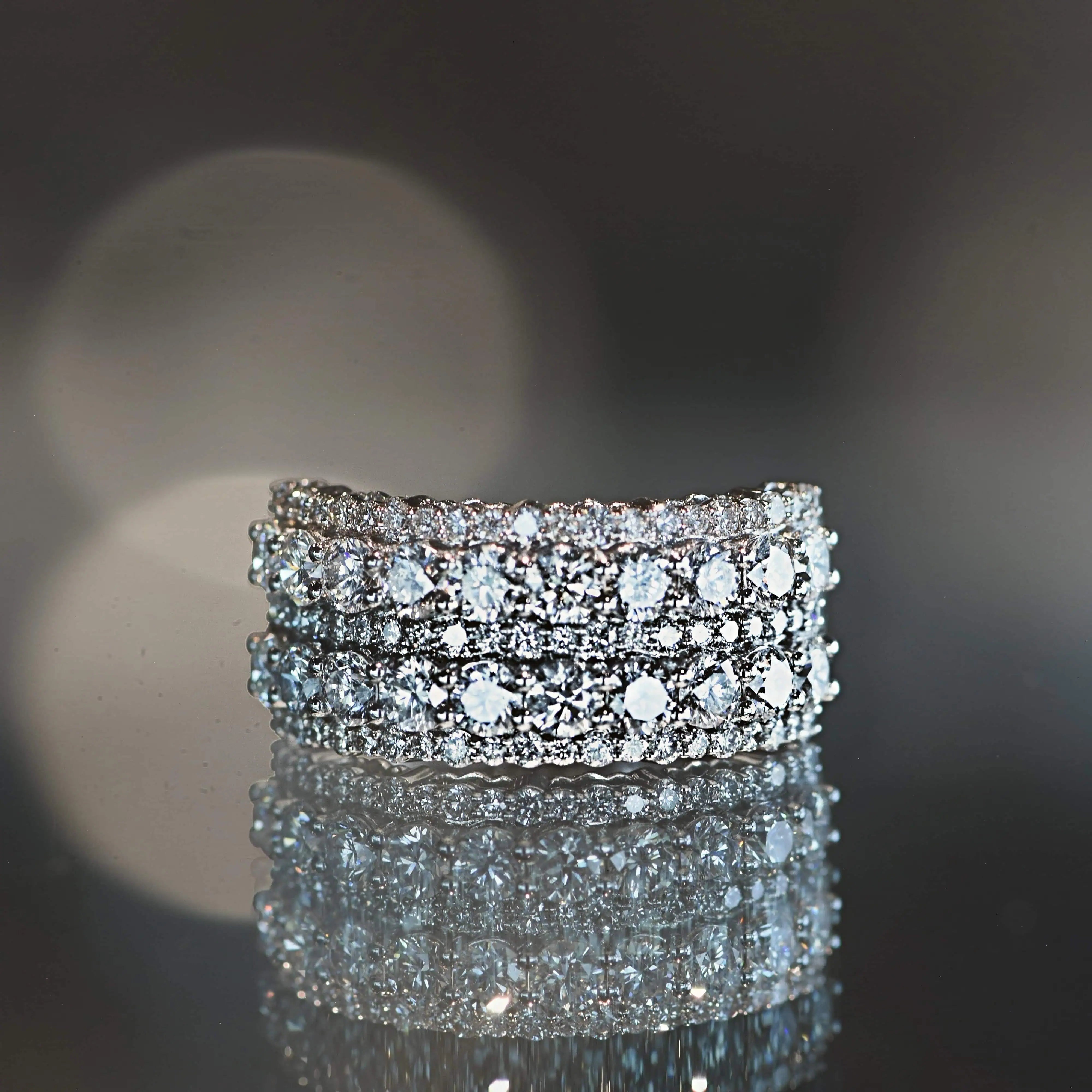 Close-up of a multi-row diamond band ring featuring round brilliant diamonds set in white metal, displayed on a reflective surface.