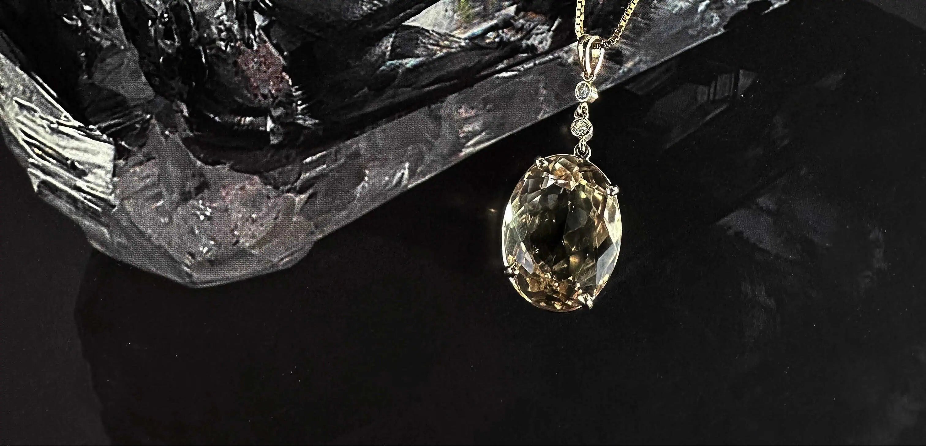 Gold pendant necklace featuring a large faceted light brown gemstone with small diamond accents, displayed against a dark textured background.