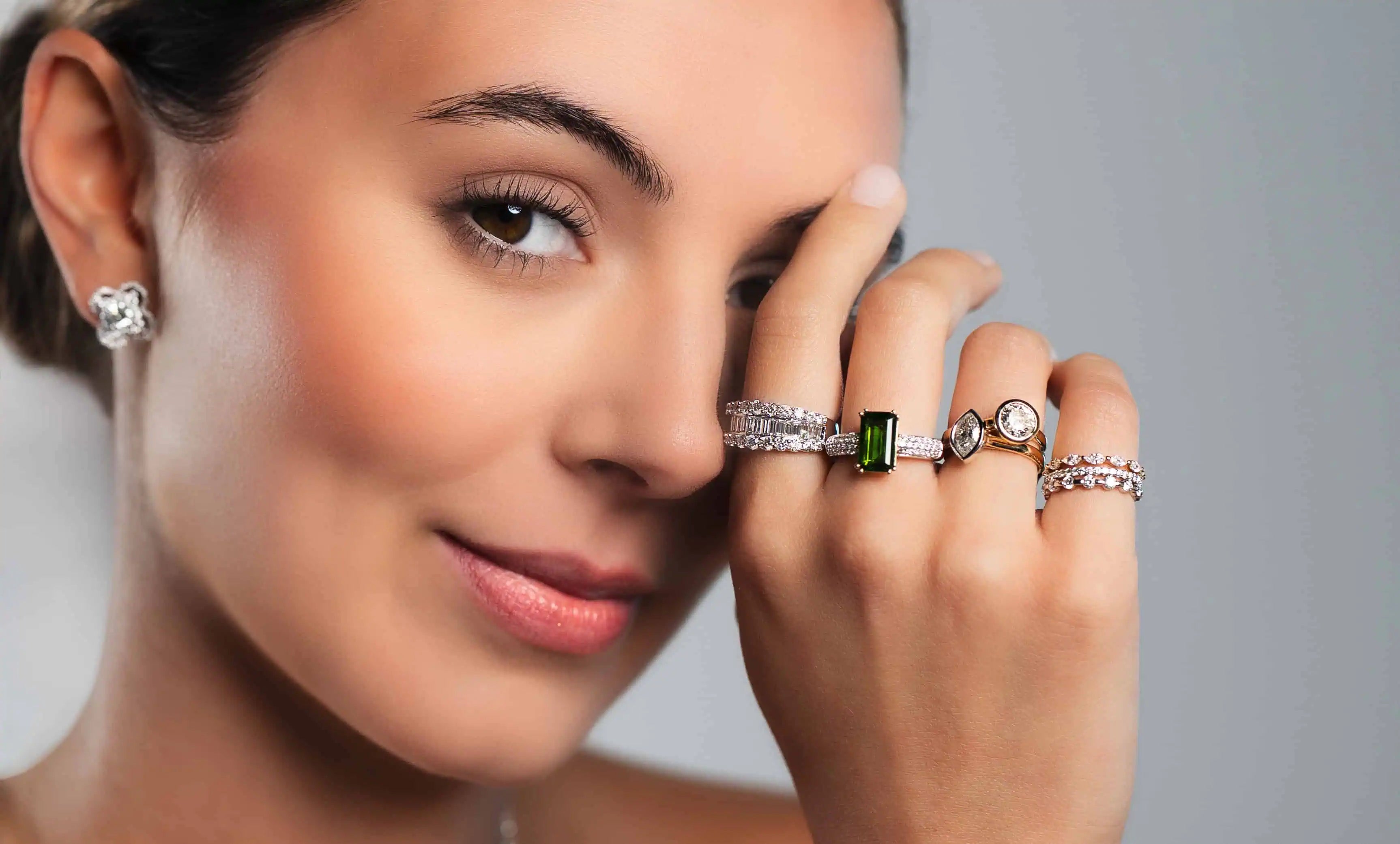 Elegant close-up of a woman wearing multiple diamond rings, including a green emerald-cut gemstone ring, stacked bands, and a diamond stud earring against a neutral background.