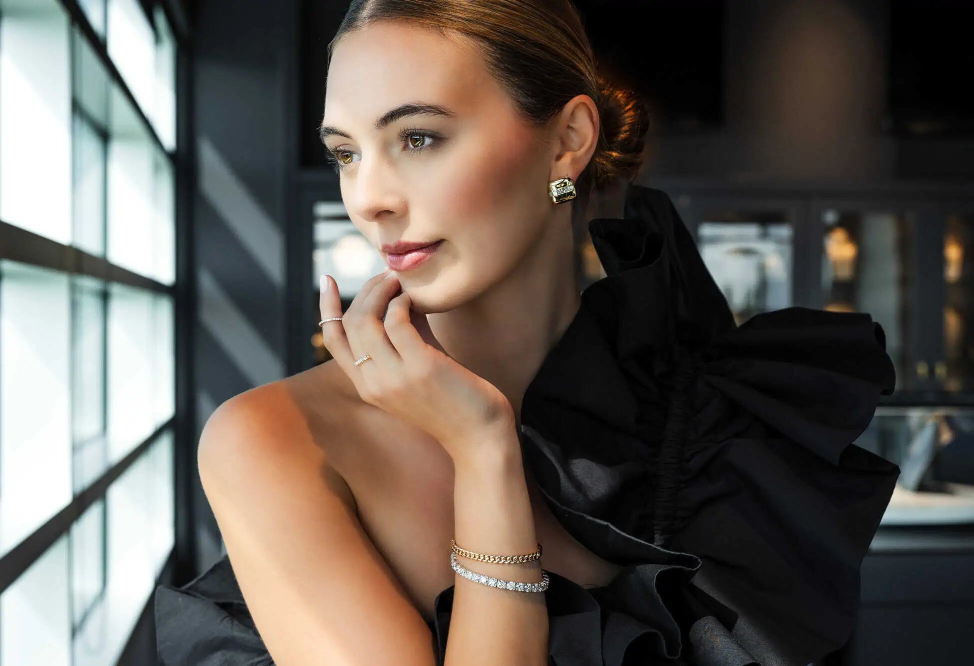 Elegant woman wearing gold and diamond jewelry—including stud earrings, stacked bracelets, and delicate rings—posed in a luxury jewelry showroom with soft natural lighting.