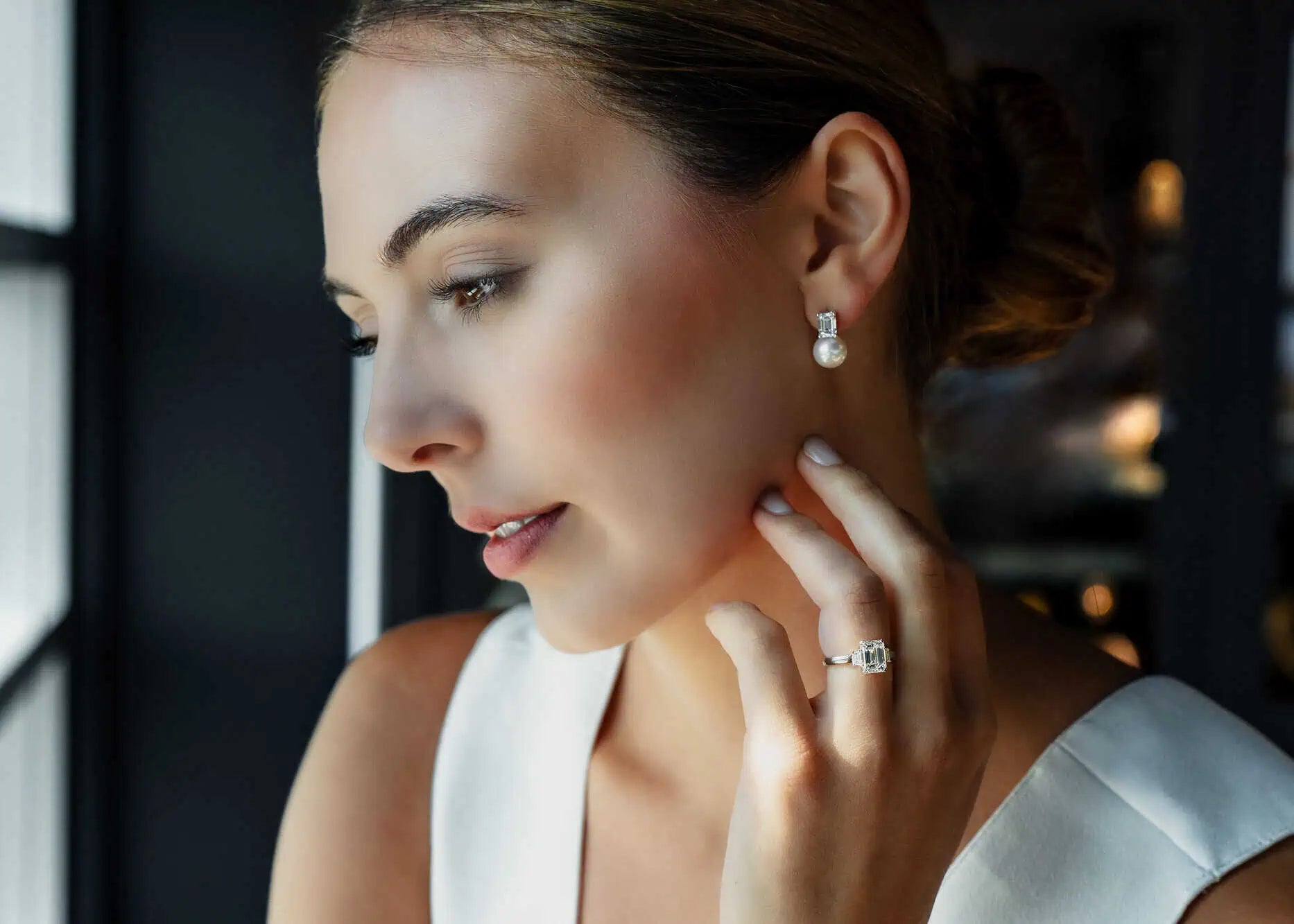 Elegant woman wearing pearl drop earrings and an emerald-cut diamond ring, posed in soft natural light inside a luxury jewelry showroom.