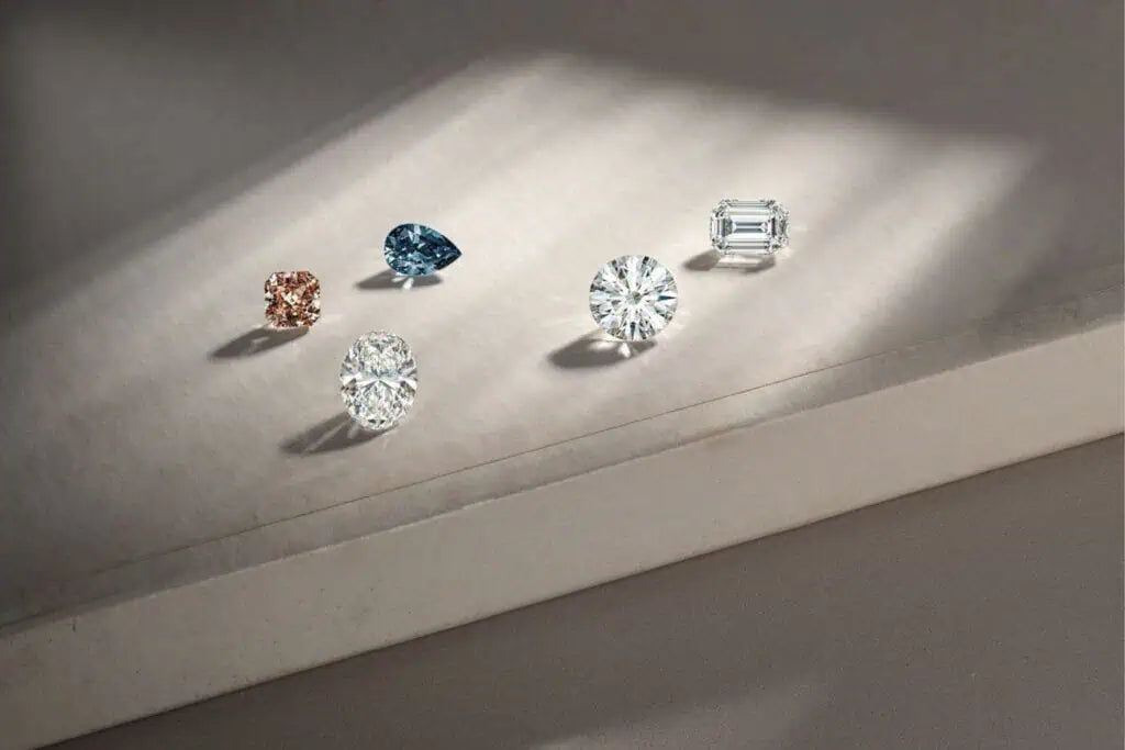 Collection of loose diamonds in various cuts and colors—including round, oval, pear, emerald, and cushion—displayed on a softly lit neutral surface.