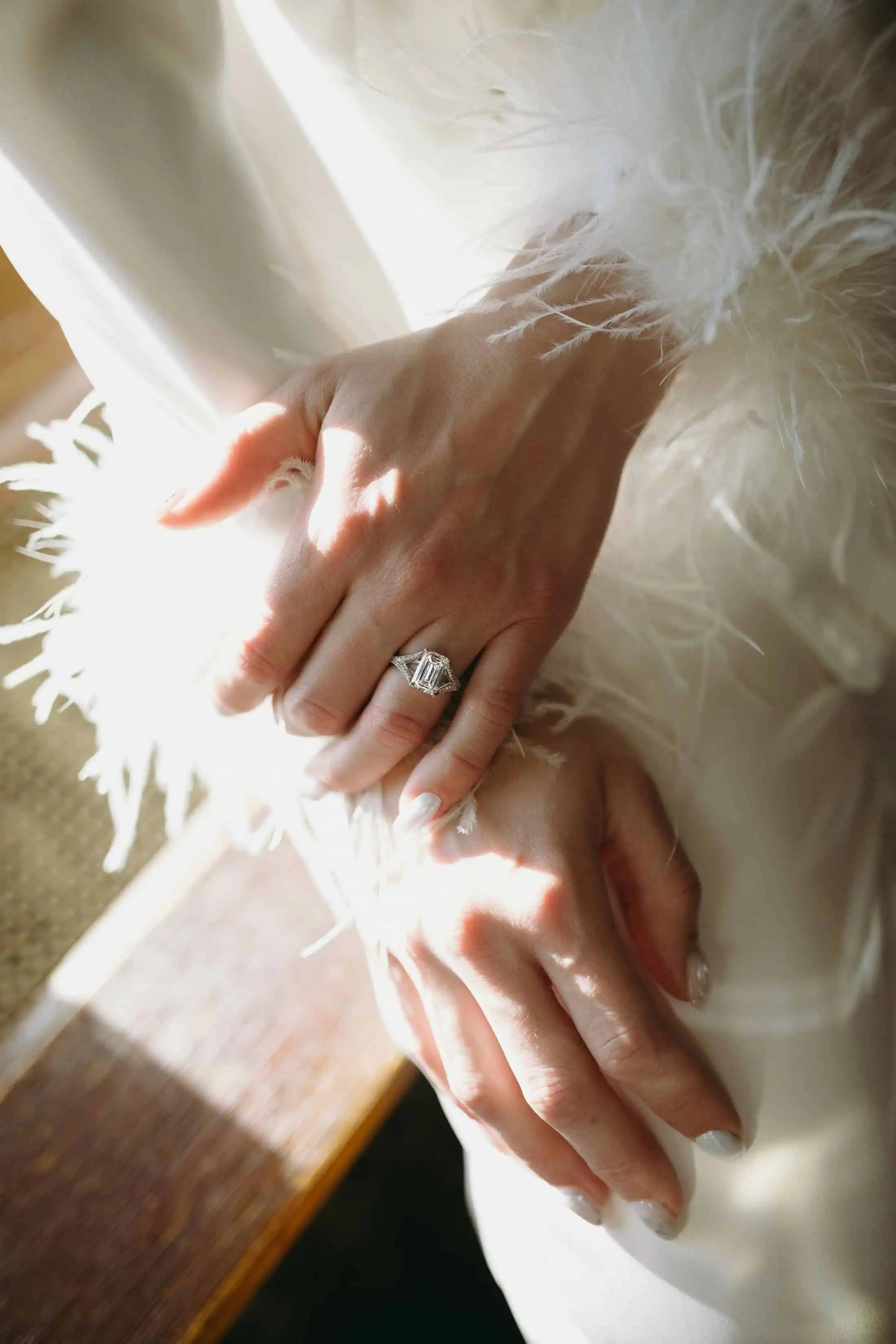Close-up of a bride’s hands wearing an emerald-cut diamond engagement ring, highlighted by soft natural light and feather-trimmed bridal sleeves.