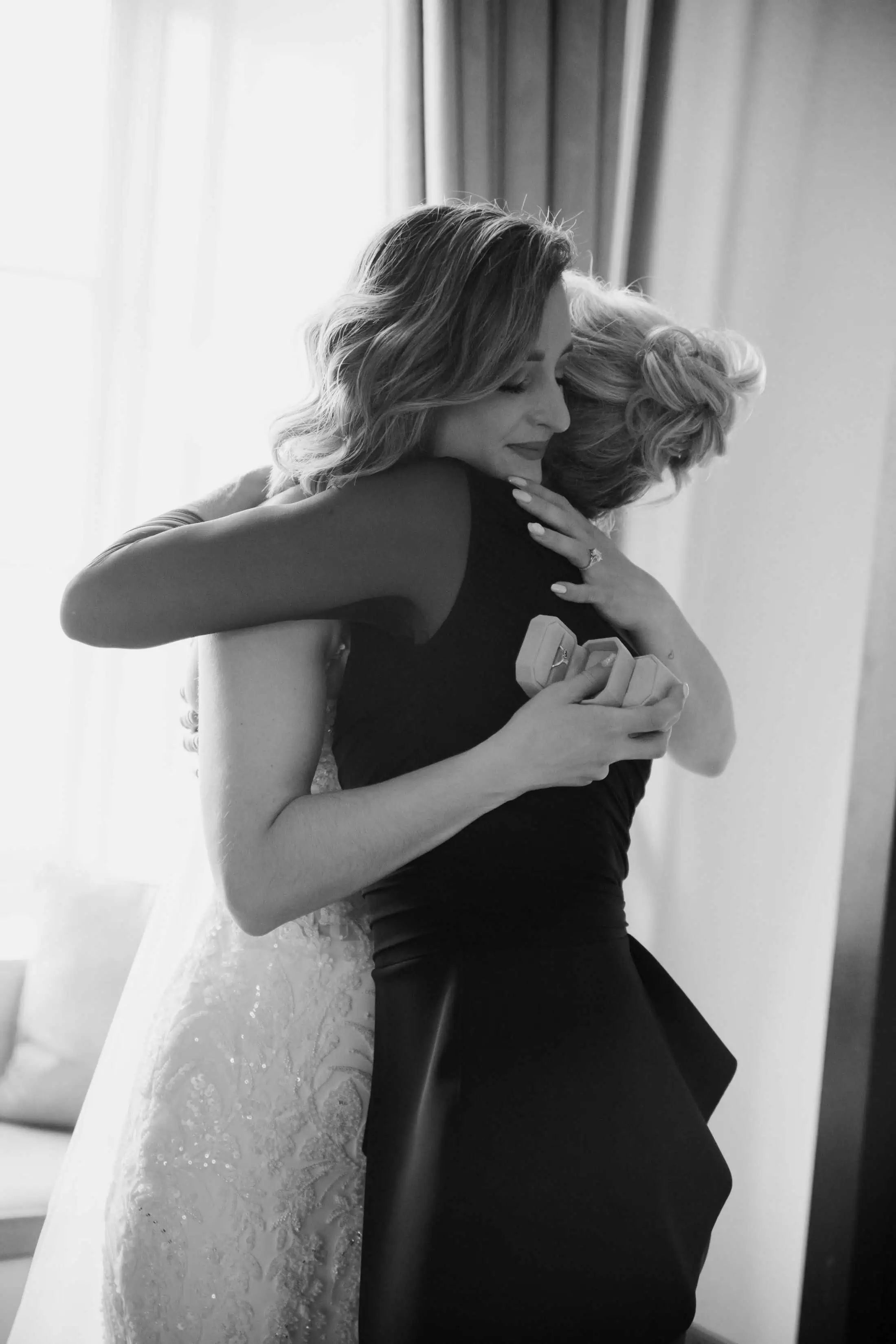 Black-and-white photo of a bride hugging a loved one while holding a ring box, capturing an emotional wedding moment.