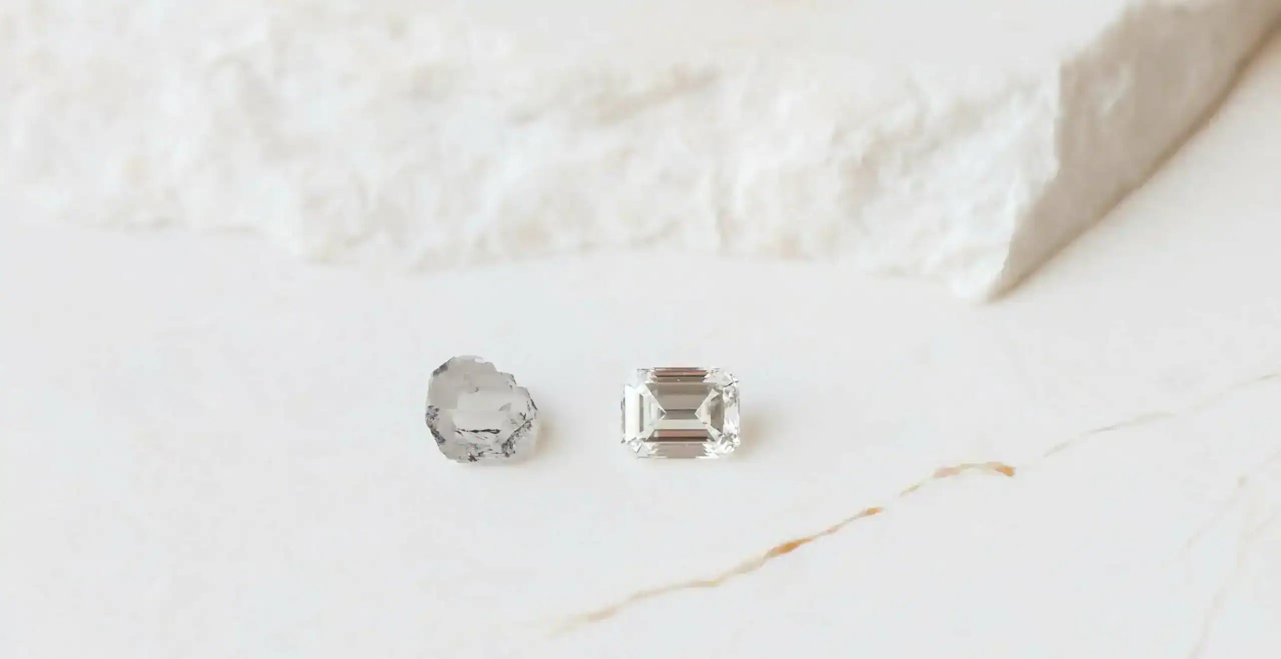 Comparison of a rough uncut diamond and a polished emerald-cut diamond placed on a light stone surface, illustrating the transformation from raw gemstone to finished cut.