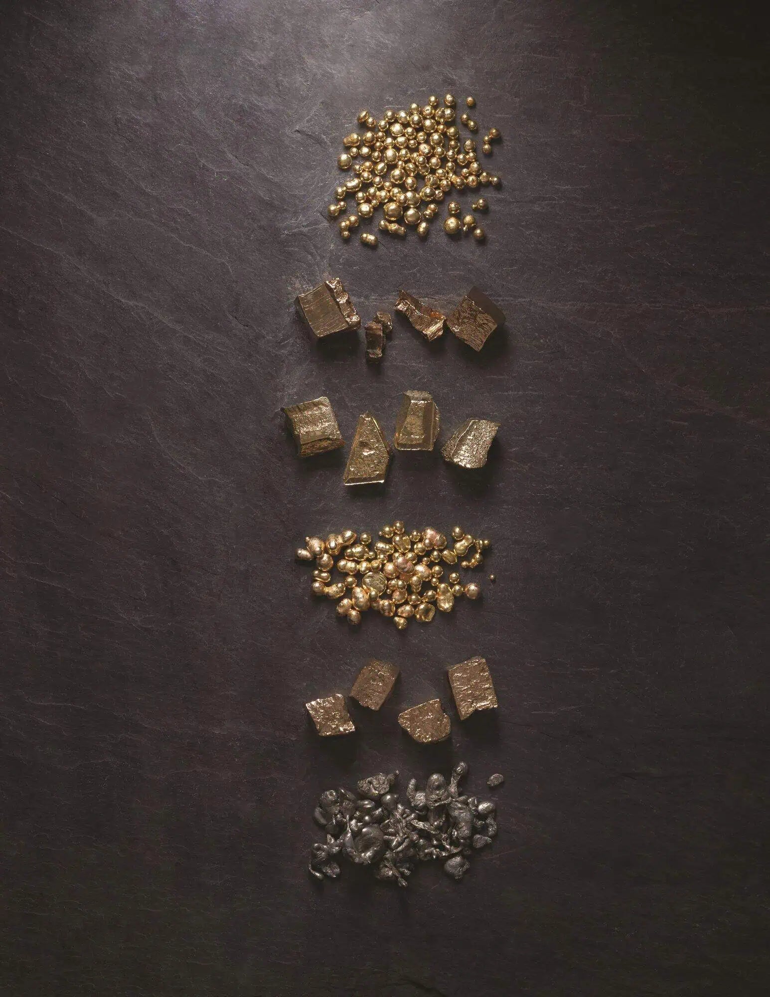 Arrangement of raw gold nuggets, gold granules, and precious metal pieces displayed on a dark stone surface, illustrating stages of materials used in fine jewelry creation.