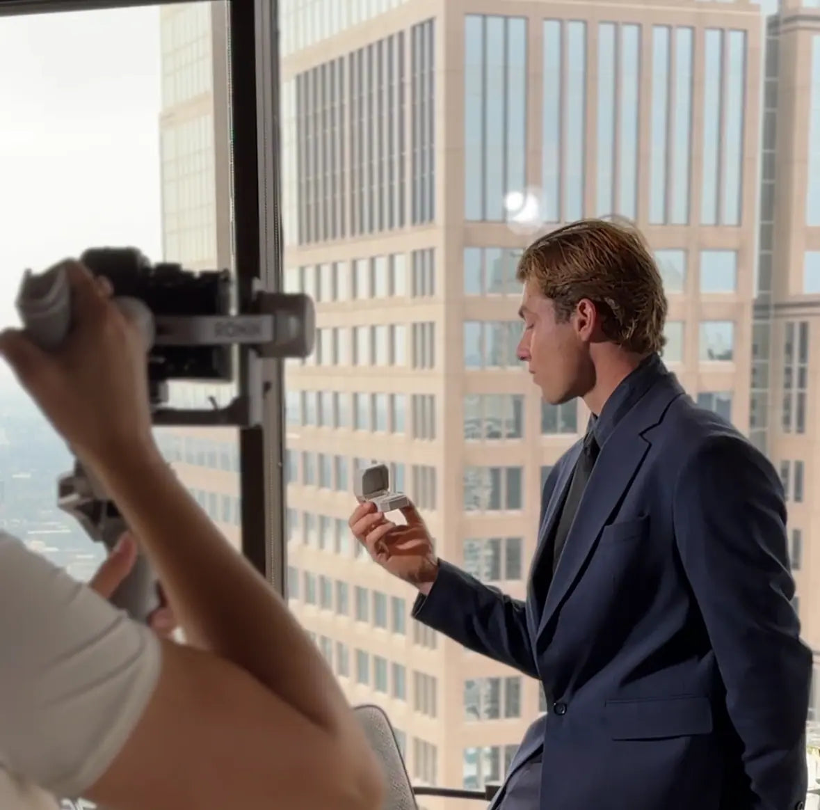Behind-the-scenes shot of a man in a suit holding an open ring box by a window overlooking city buildings, while a photographer captures the moment.