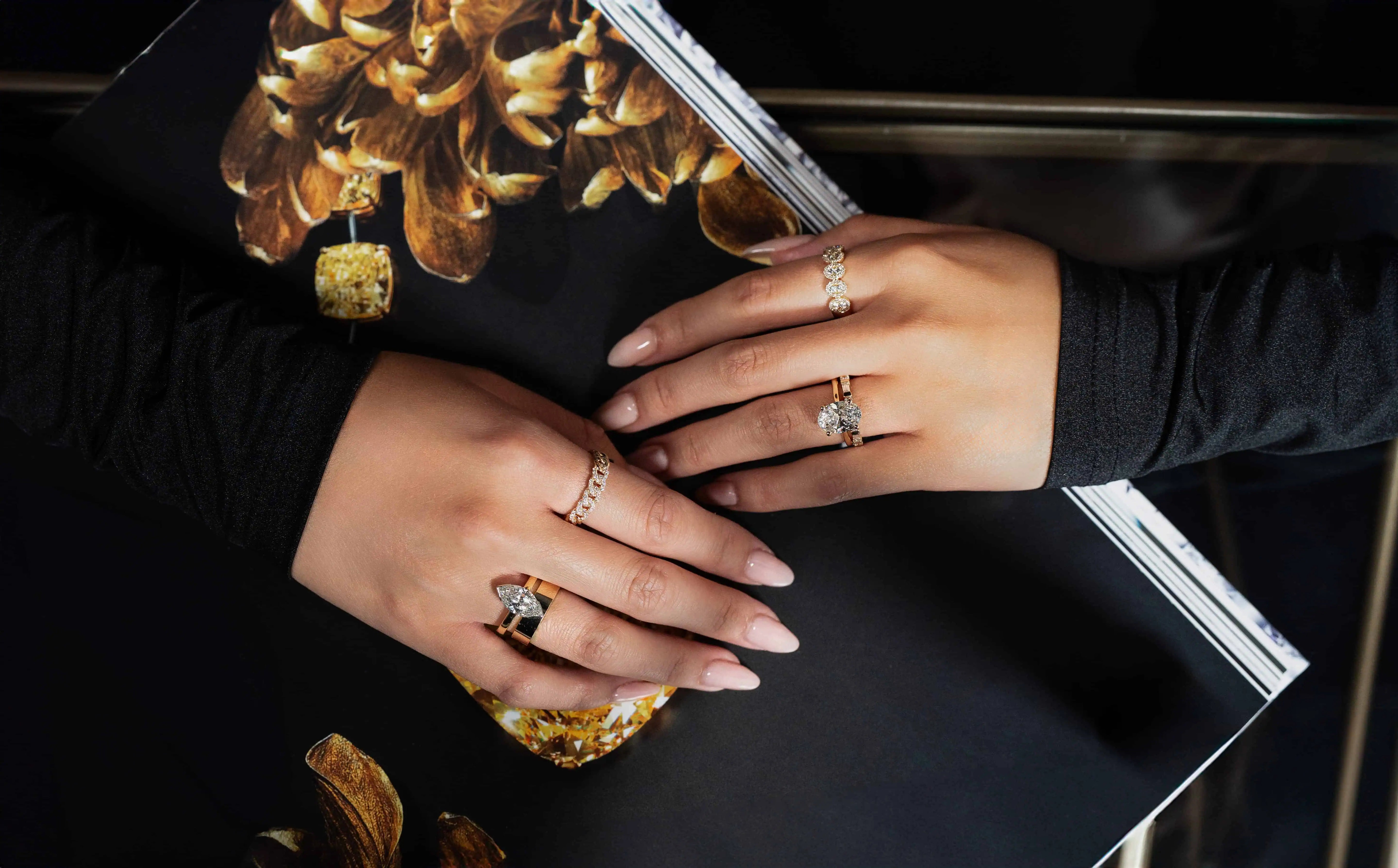 Manicured hands wearing multiple gold and diamond rings resting on a magazine featuring gemstone imagery, highlighting layered fine jewelry styling and luxury design.