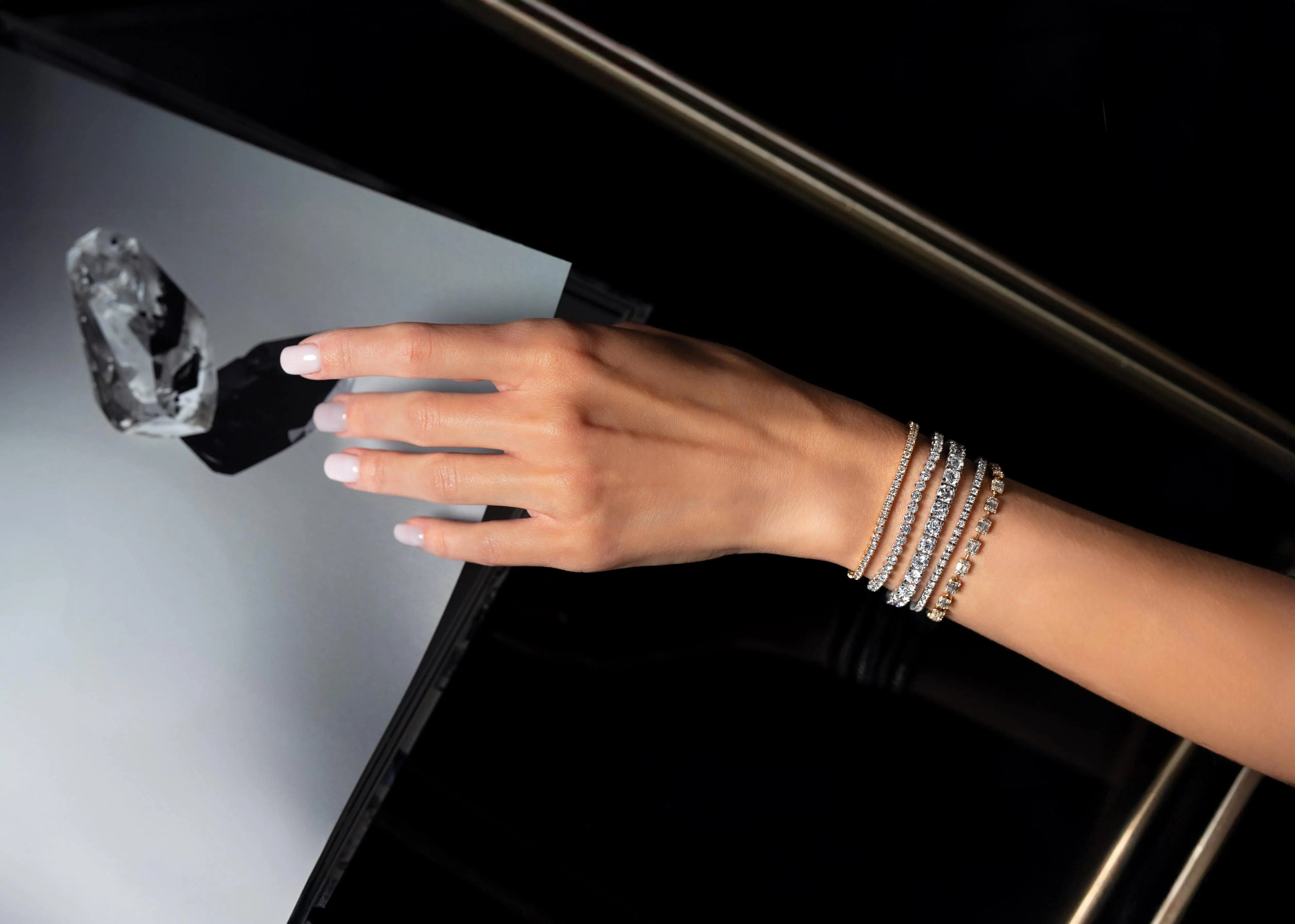 Close-up of a hand wearing stacked diamond bracelets reaching toward a large loose diamond displayed on a reflective surface.