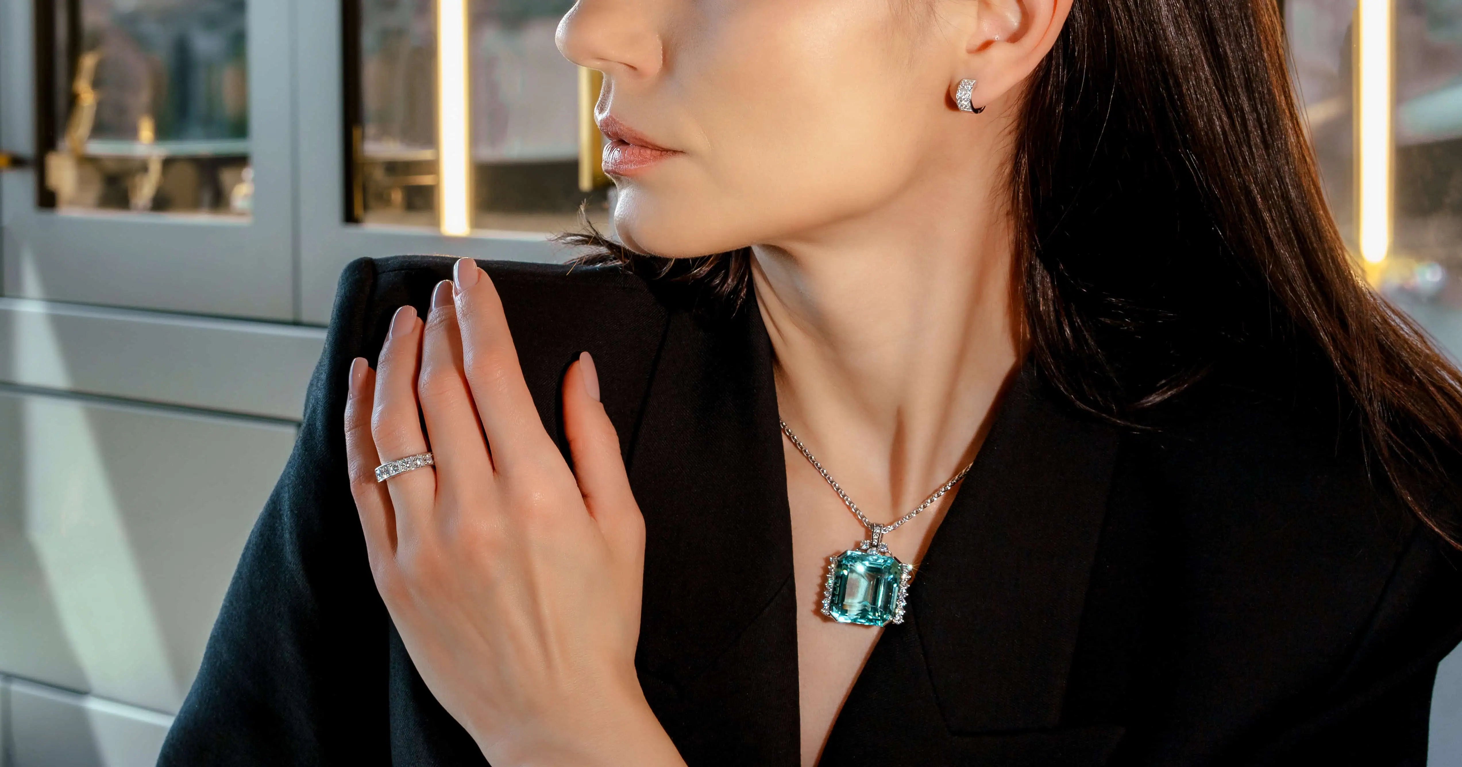 Elegant woman wearing emerald-cut green gemstone pendant necklace with diamond halo, matching diamond stud earrings, and a diamond band ring, dressed in a black blazer in a luxury jewelry showroom setting.