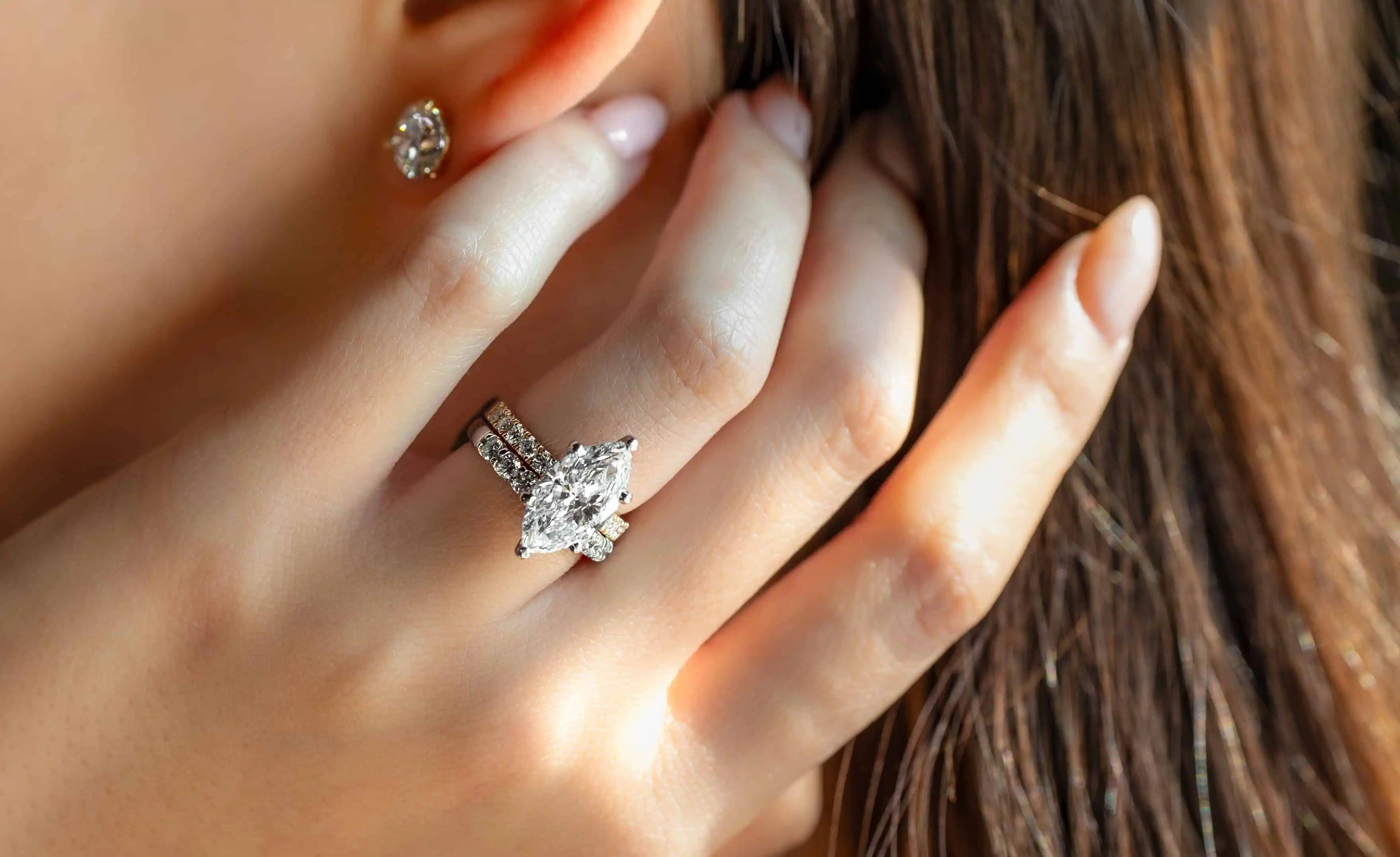 Close-up of a woman wearing a marquise-cut diamond engagement ring with a diamond band, paired with a matching diamond stud earring.